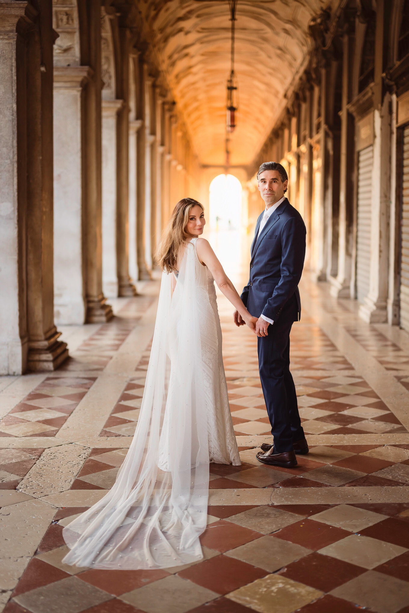 A romantic couple holding hands in a sunlit Venetian arcade, surrounded by historic architecture and warm golden light.