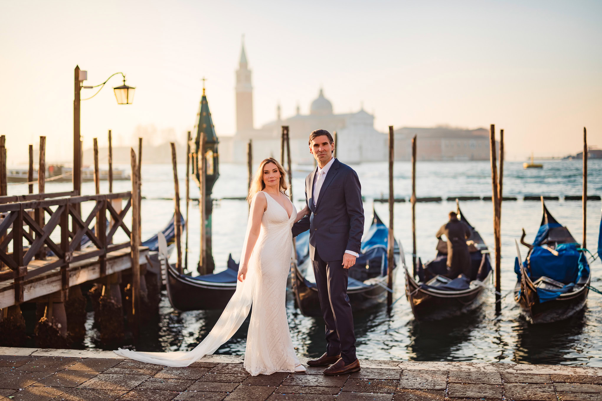 Romantic couple standing by gondolas in Venice at sunset, with warm light and historic cityscape in the background.