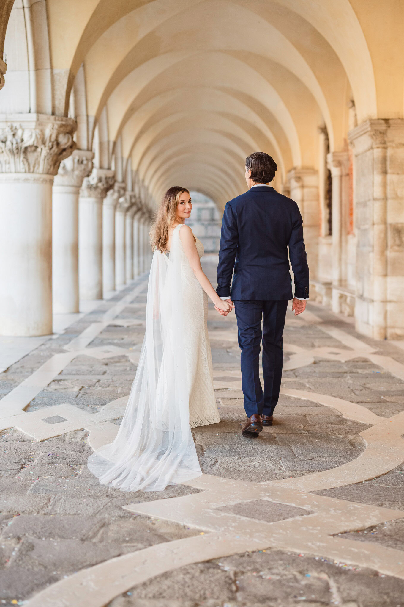 A couple holding hands and walking through a historic Venetian arcade bathed in soft, romantic light.