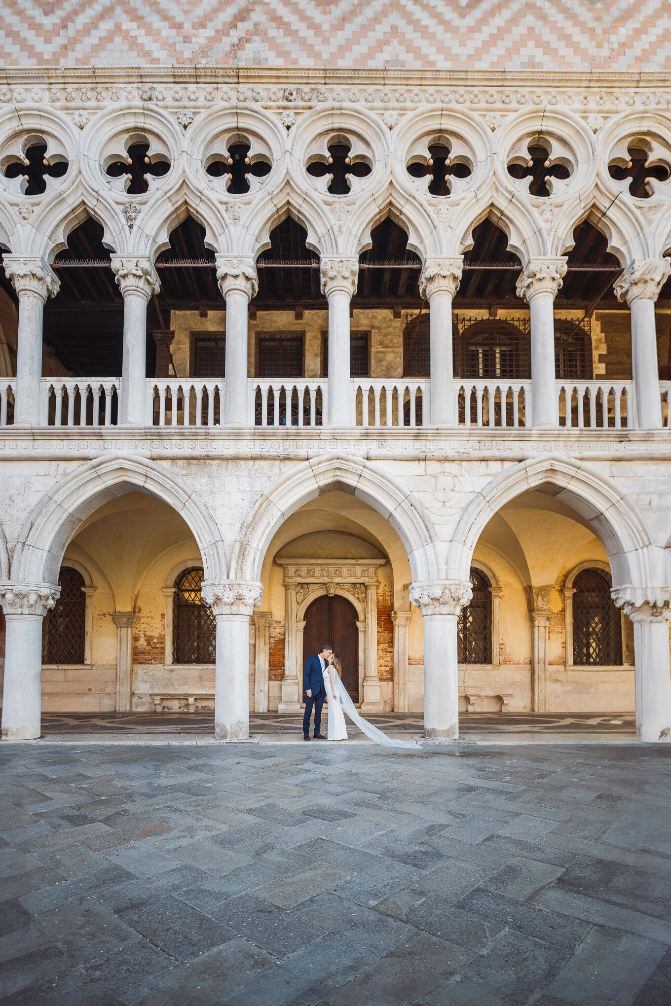 A couple in elegant wedding attire sharing a romantic moment beneath historic Venetian arches at sunset.