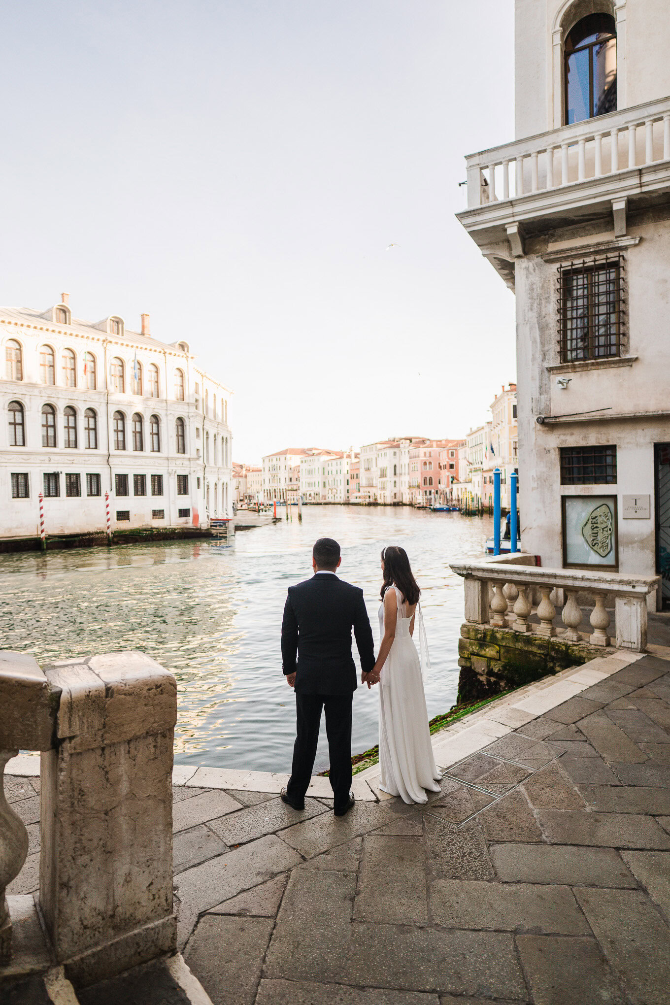 A couple in elegant attire stands by the canal in Venice, bathed in soft natural light, creating a romantic and timeless.