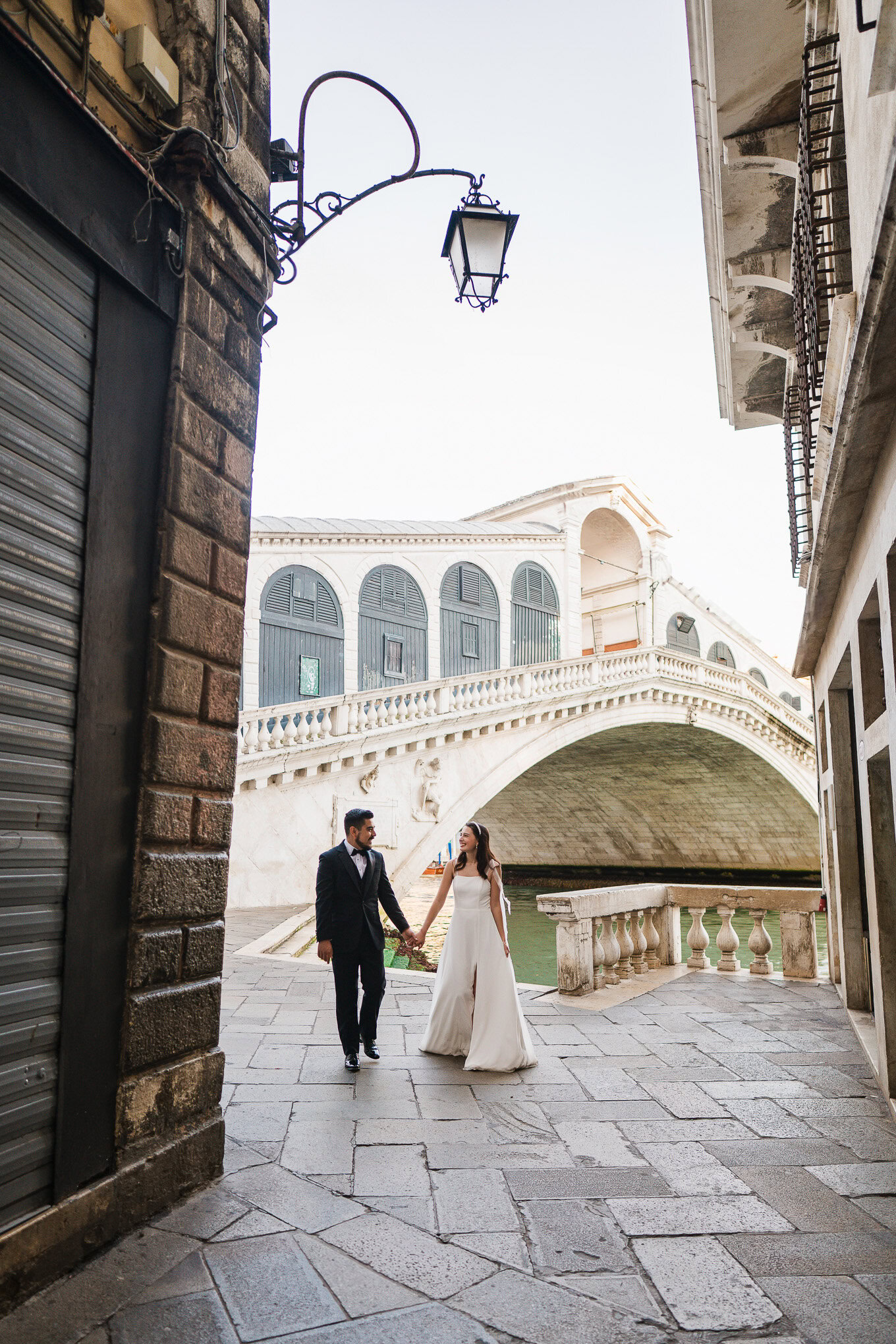 A couple holding hands and strolling along a Venetian canal under soft daylight, exuding romance and serenity.