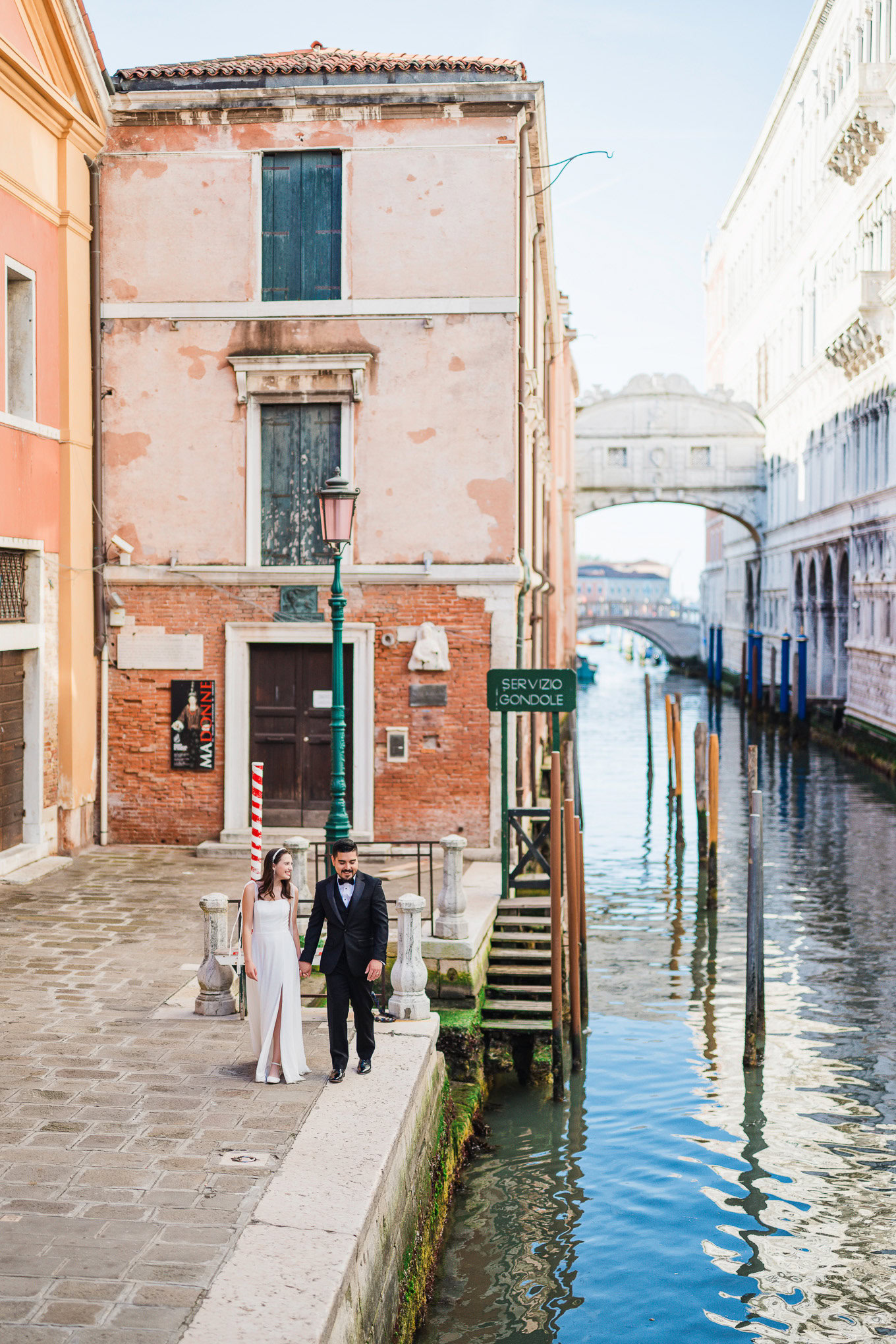 A couple walking hand in hand along a romantic Venice canal bathed in soft daylight.
