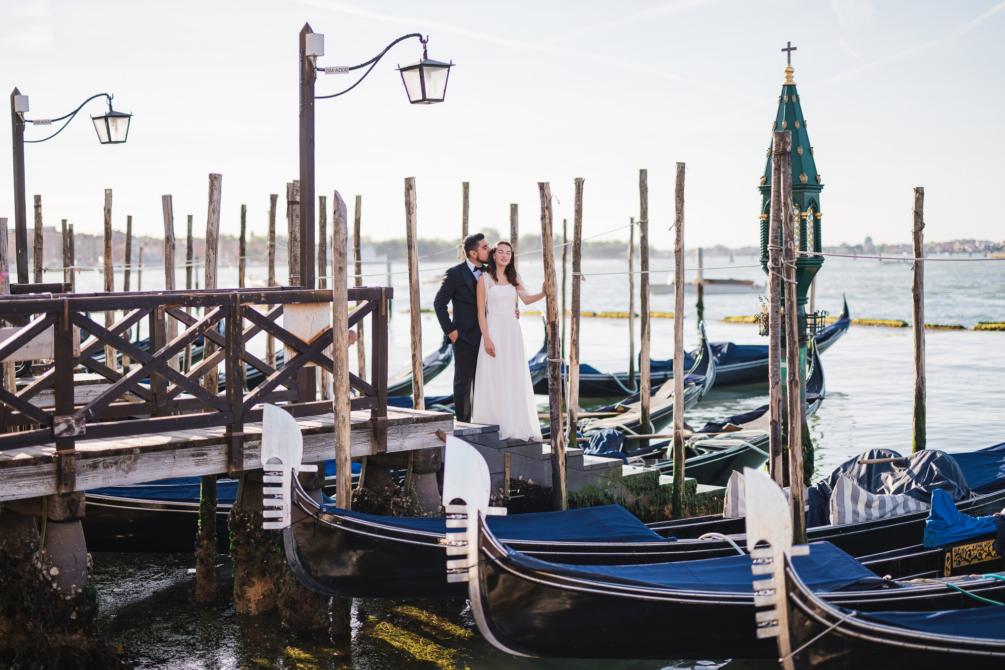 A couple standing on a wooden dock by gondolas in Venice, bathed in soft natural light, creating a romantic and serene a.