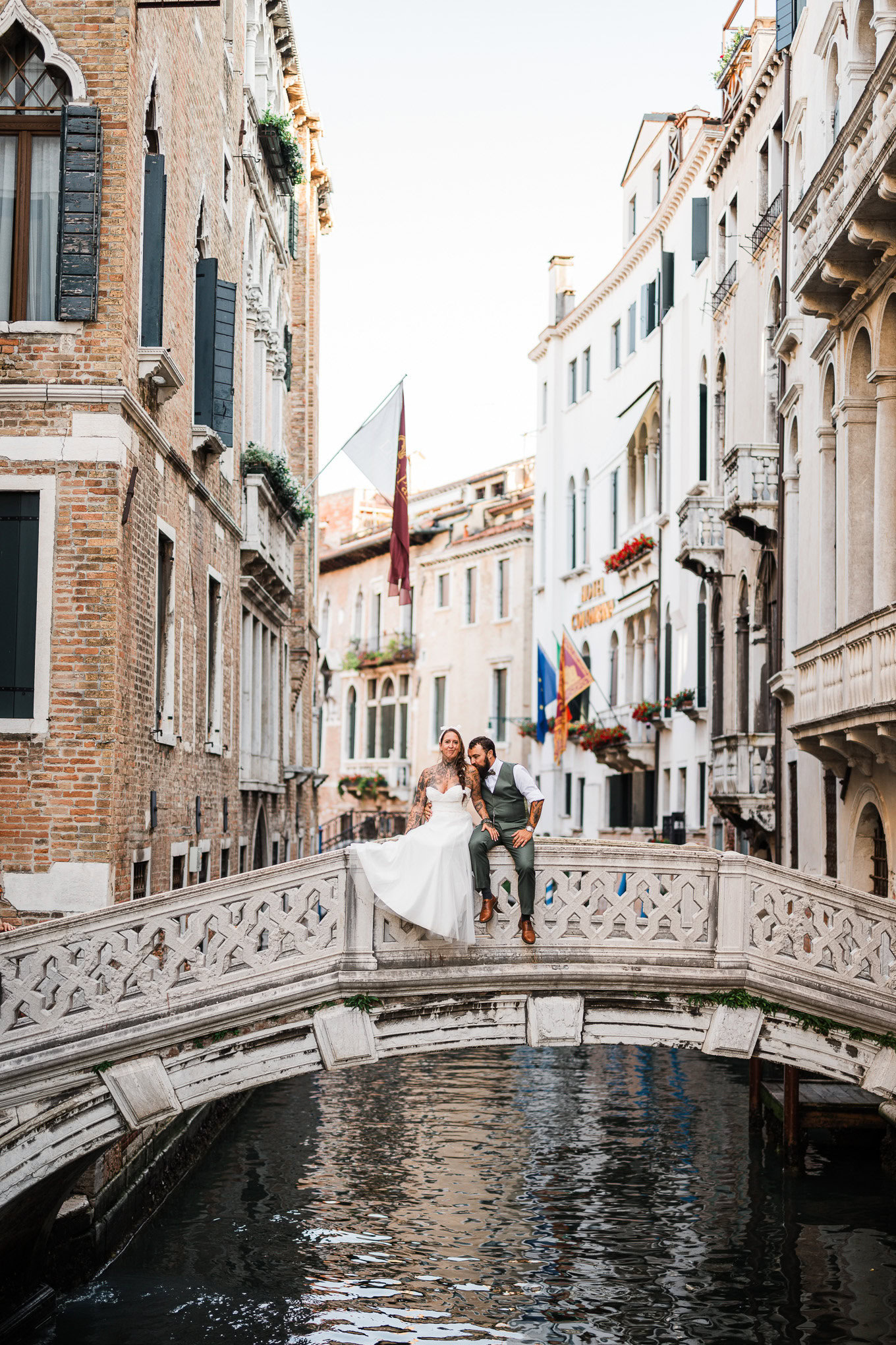 A couple sitting on a small bridge over a canal in Venice, surrounded by historic buildings, under soft natural light, e.
