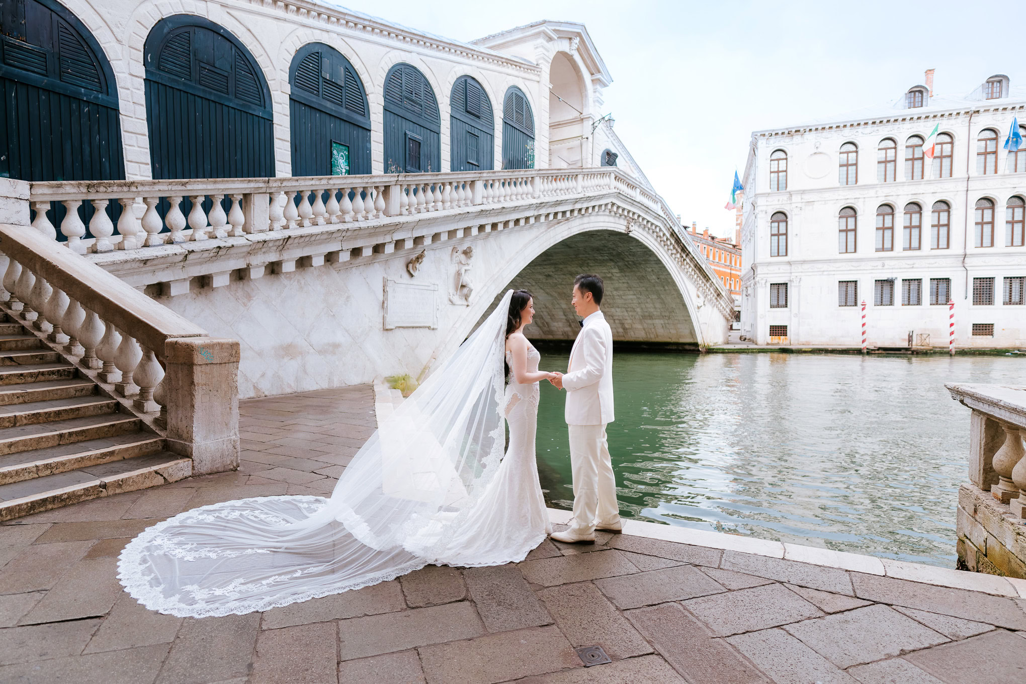 A couple in elegant wedding attire sharing a tender moment by the canal under soft daylight in Venice.