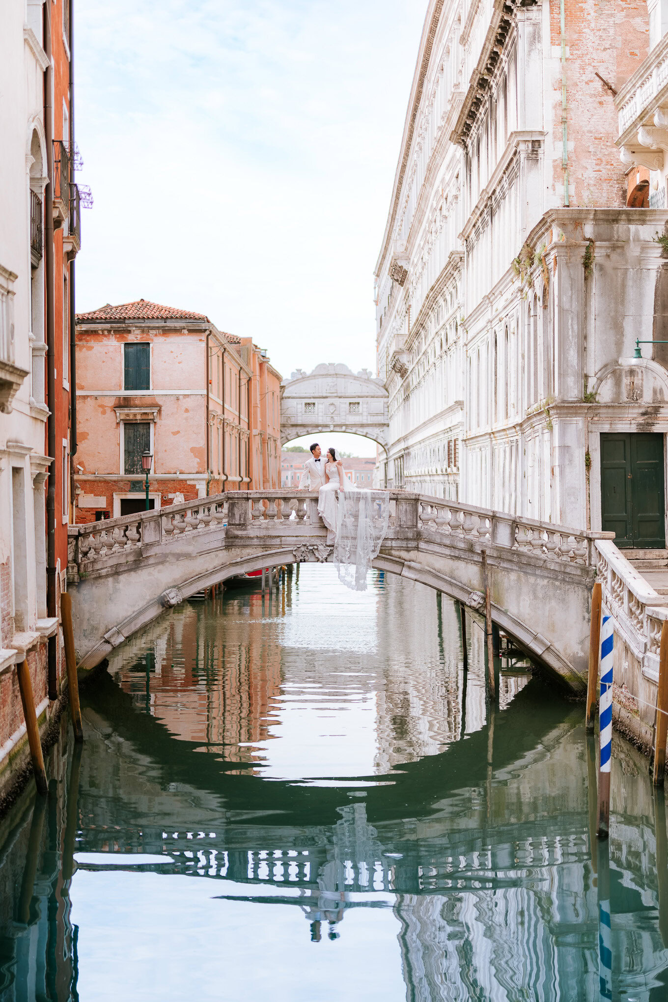 A couple standing on a small bridge over a canal in Venice, bathed in soft natural light during their honeymoon.