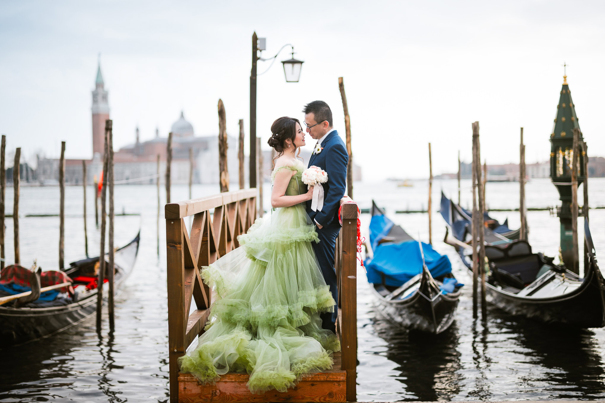 Romantic couple standing on a wooden dock in Venice with soft natural light and gondolas in the background.