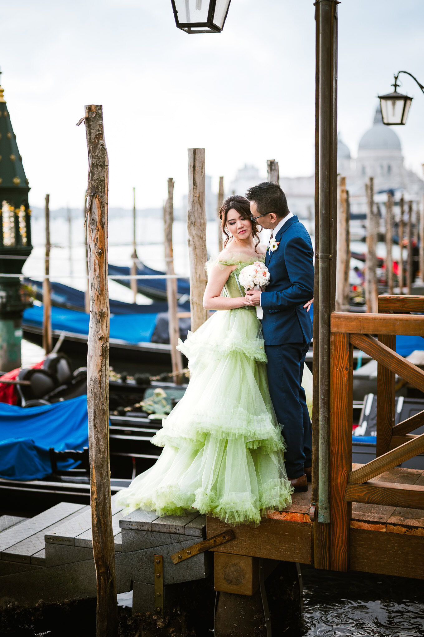 A couple in elegant attire sharing an intimate moment on a Venice dock with boats and historic architecture in the backg.