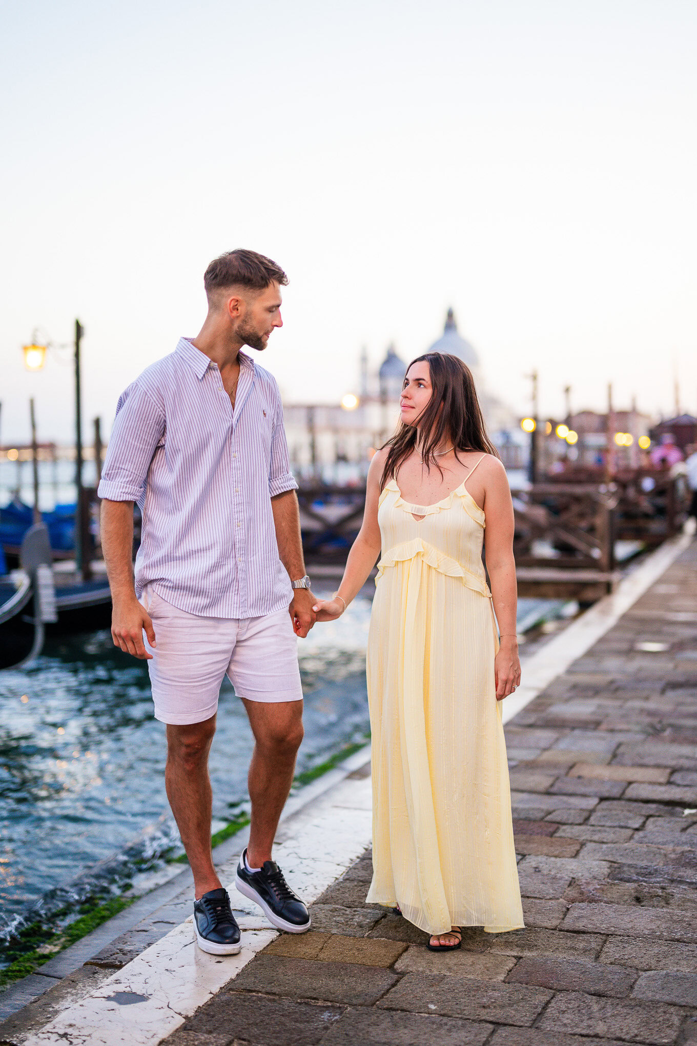 A couple holding hands walking along a Venetian canal promenade at sunset, with historic buildings and boats in the back.