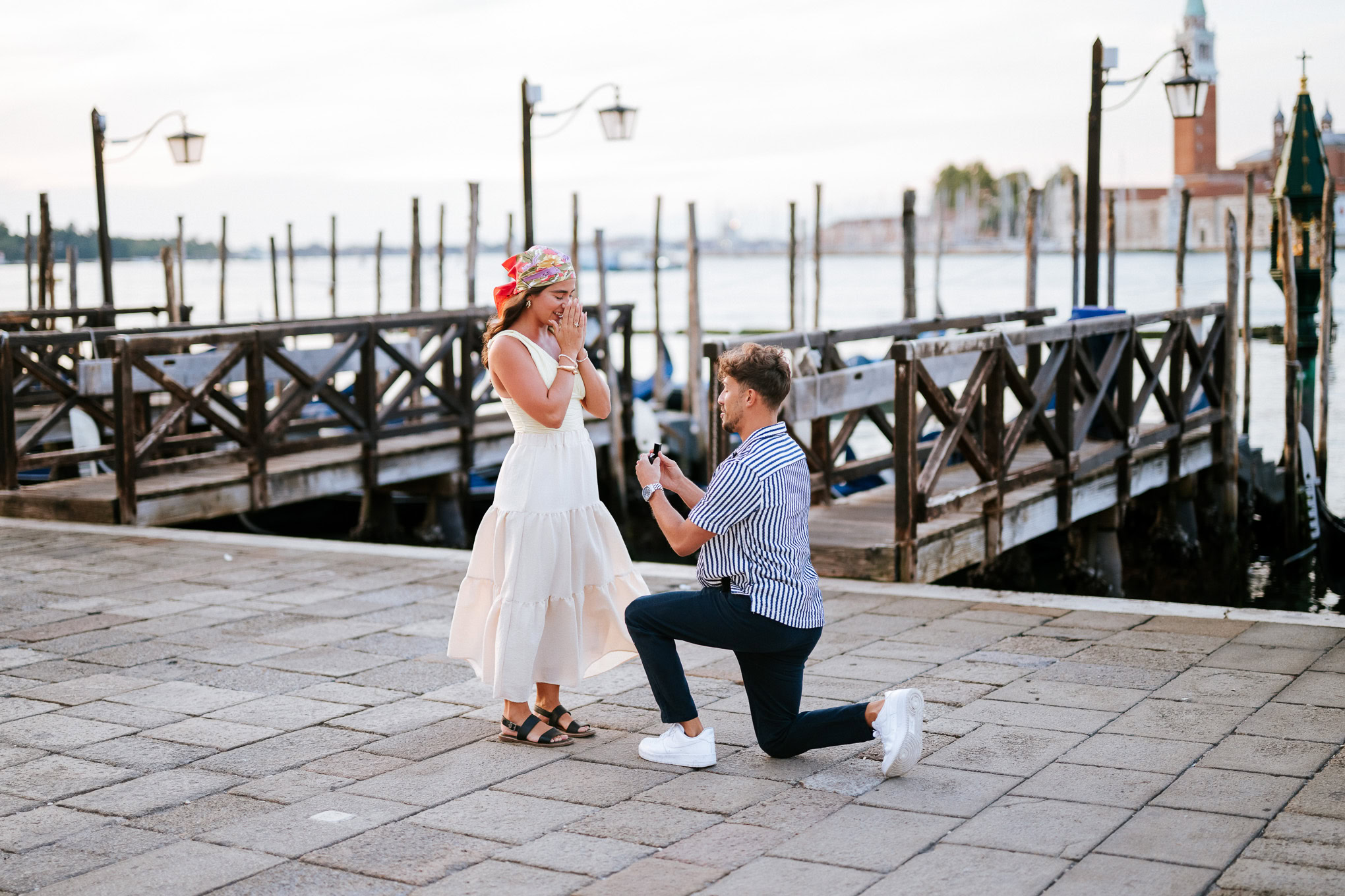 A woman in a white dress and a man proposing on a Venetian waterfront promenade with boats and lampposts in the backgrou.