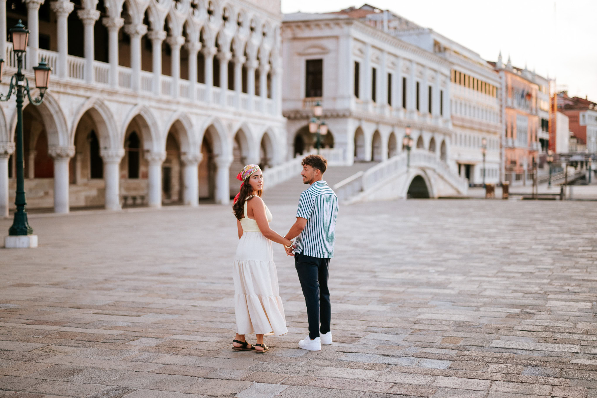 A woman and man hold hands and gaze at each other as he proposes on a historic Venetian square at sunset.
