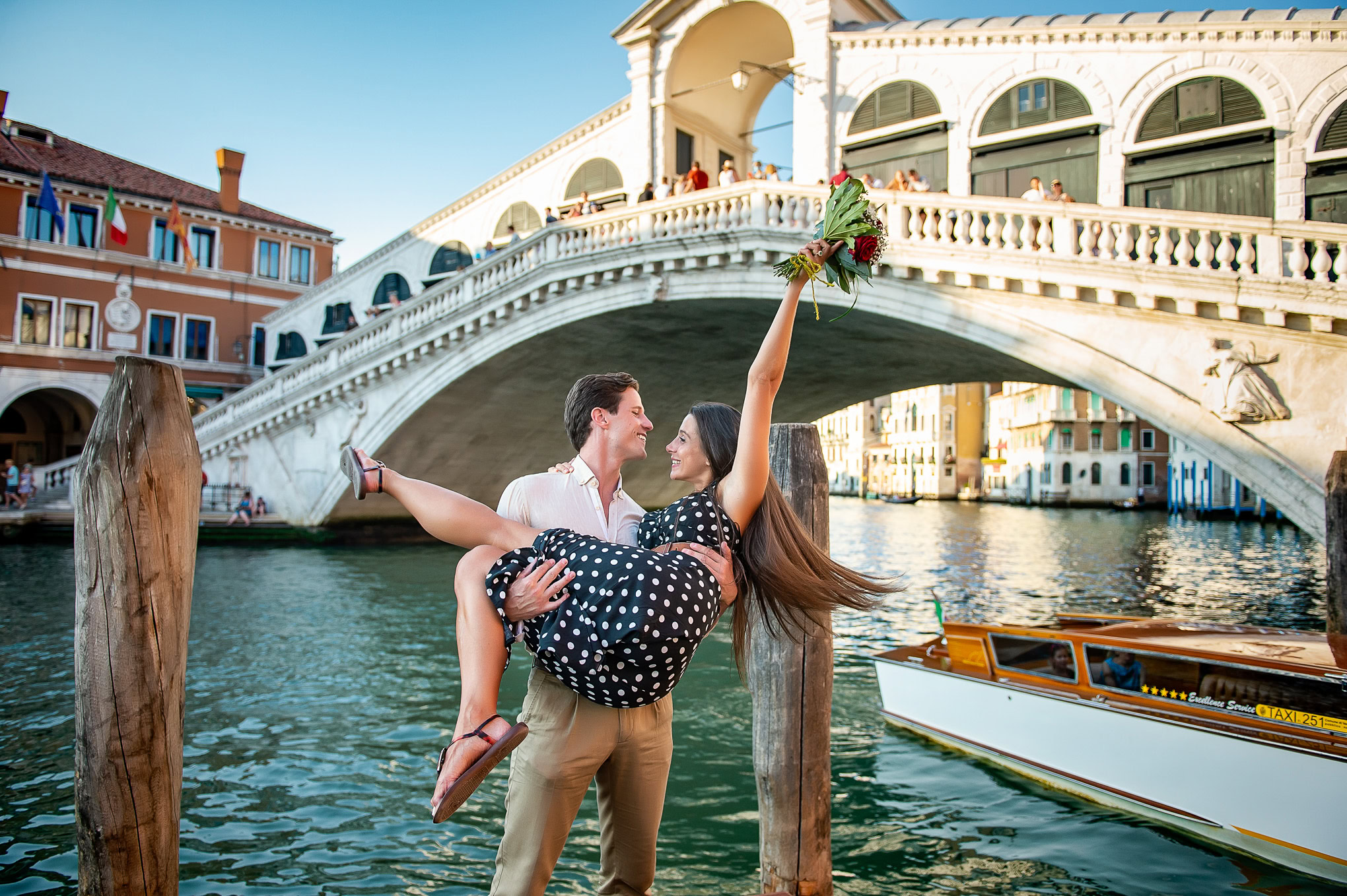 A couple celebrates a romantic proposal with a joyful embrace under Venice's iconic bridge.