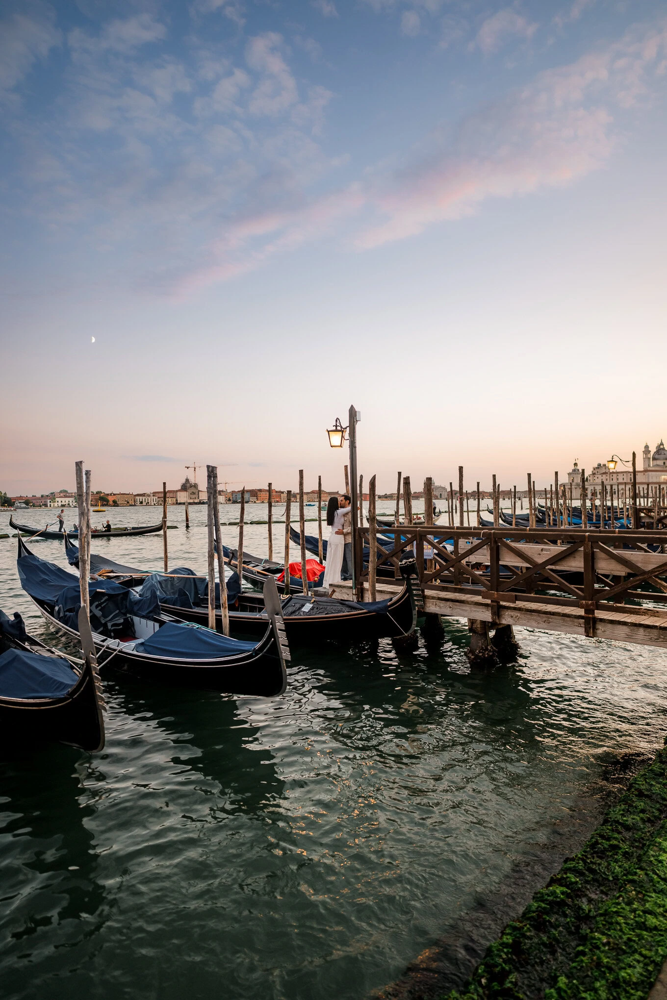 A couple shares a romantic proposal moment on a Venice dock as gondolas sway nearby at sunset.