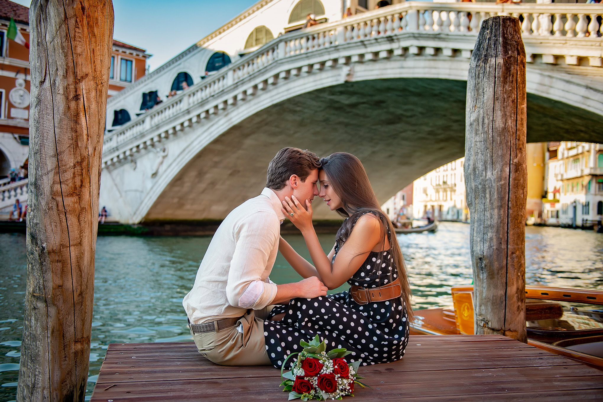 A couple sitting closely on a Venice dock with the Rialto Bridge in the background, capturing a romantic atmosphere.