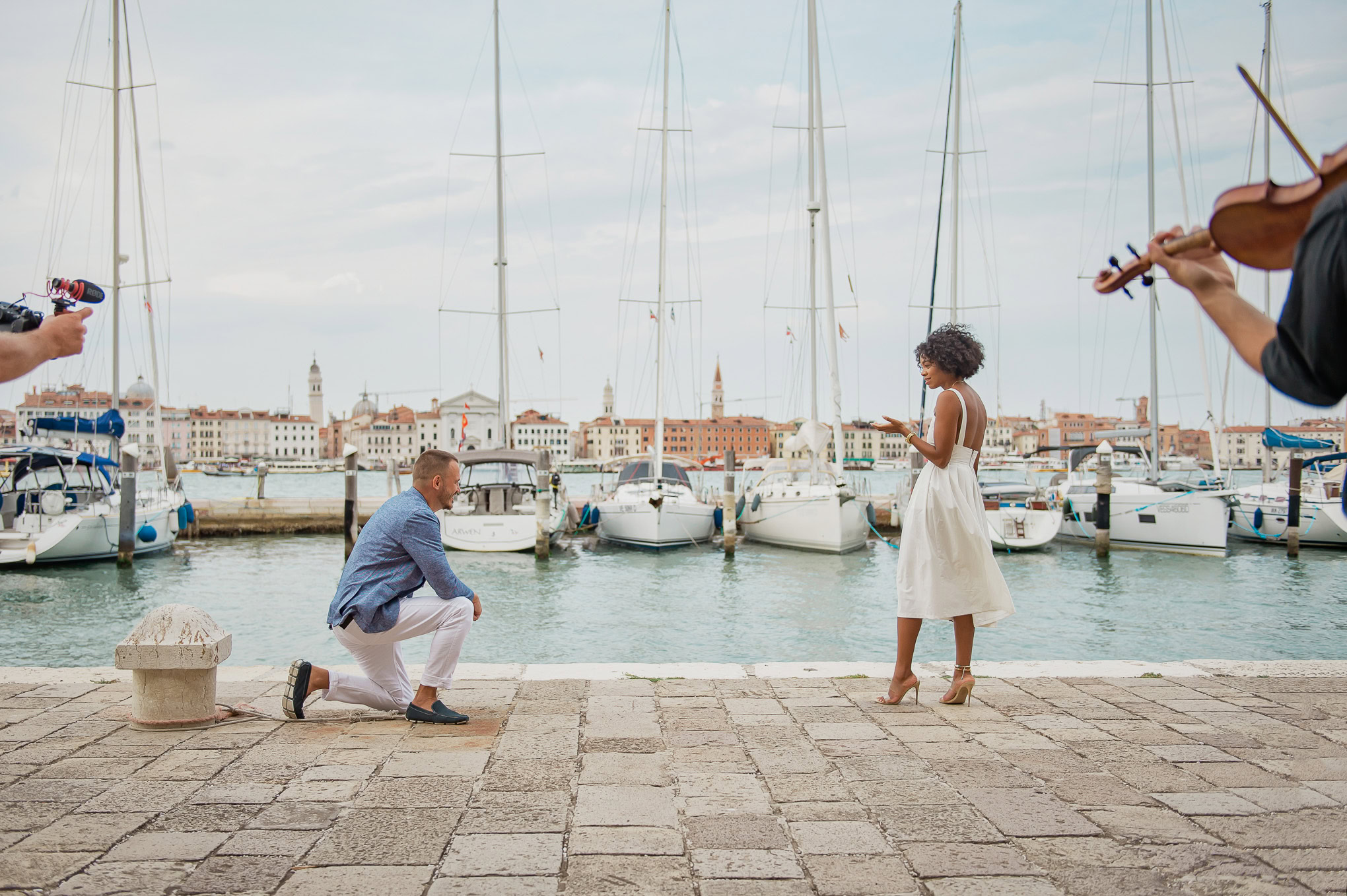 A man kneels with a ring as a woman reacts emotionally during a romantic proposal by the Venetian canal.