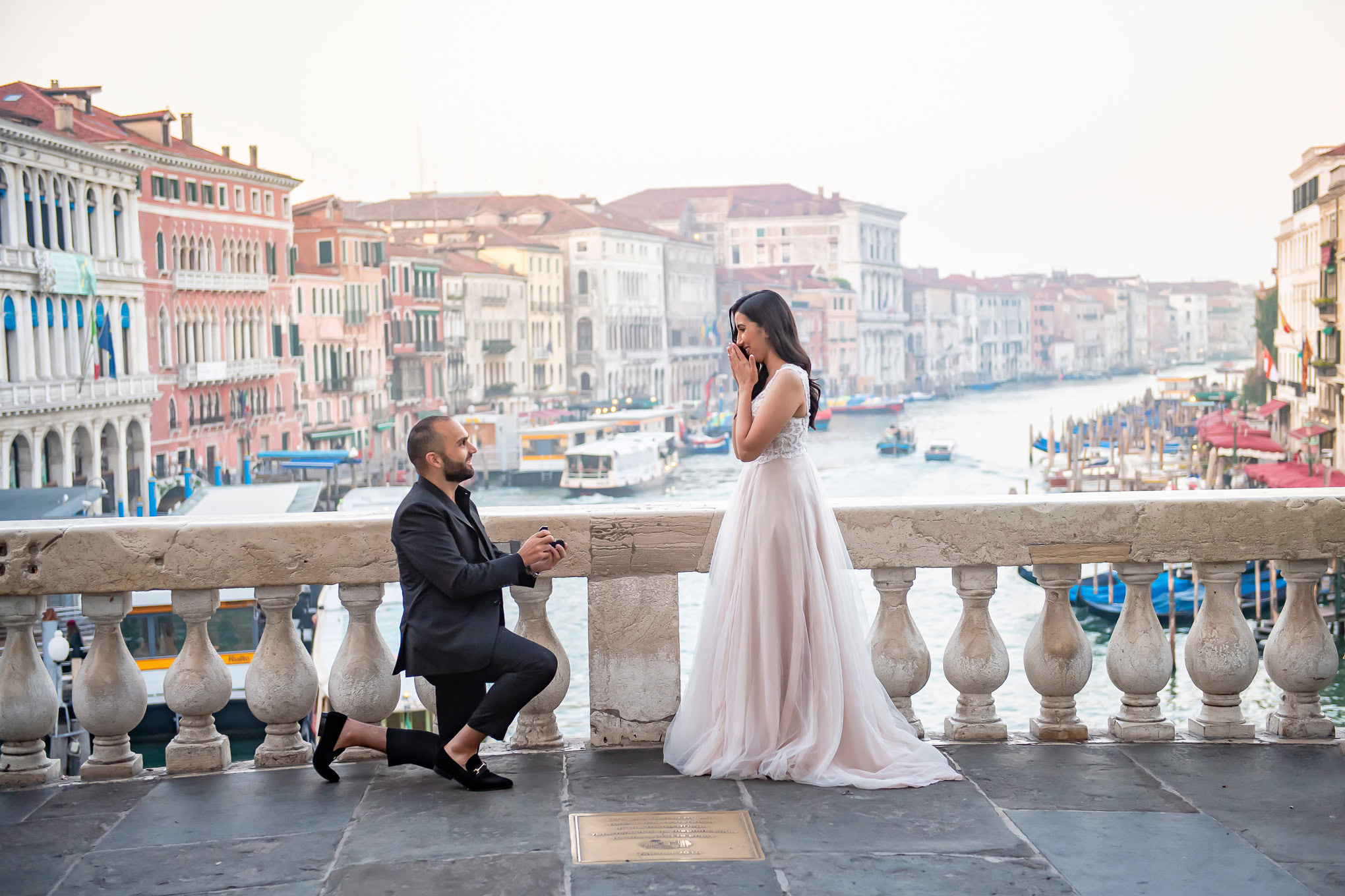 A man proposes to a woman on a Venice bridge, capturing a heartfelt moment with the canal and colorful buildings in the.