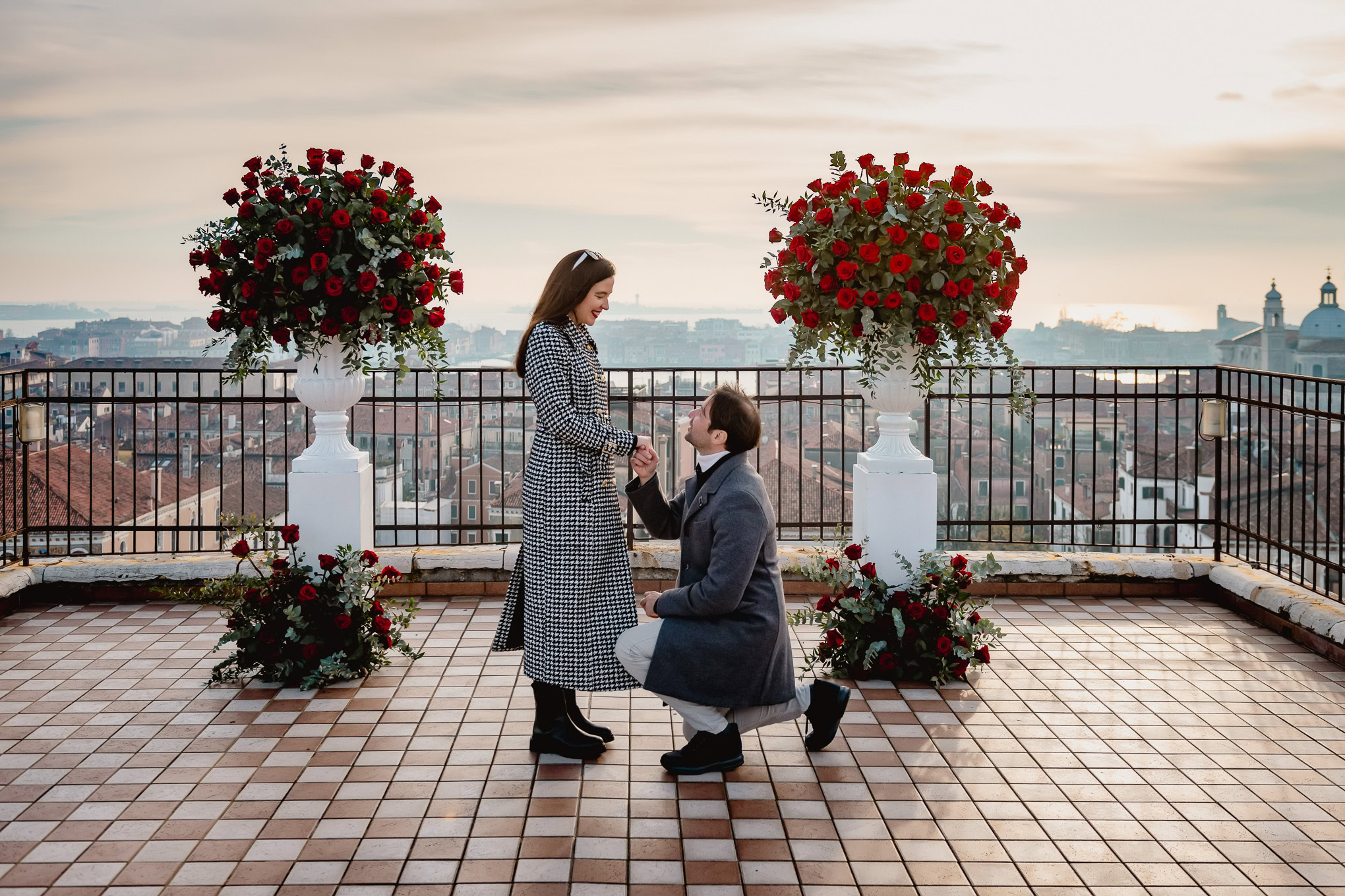A woman reacts with joy as a man proposes on a scenic Venice rooftop, surrounded by vibrant red roses and a panoramic ci.