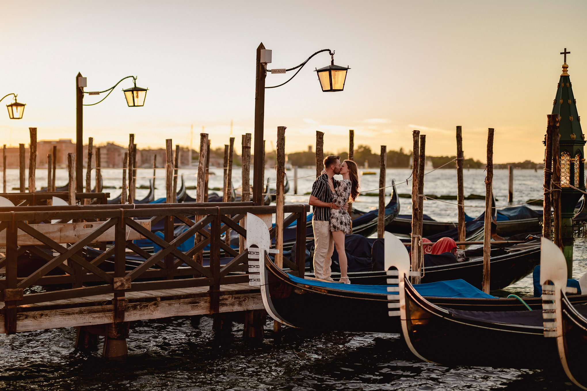A couple shares a romantic kiss on a Venice dock as the sun sets, capturing a heartfelt proposal moment by the gondolas.