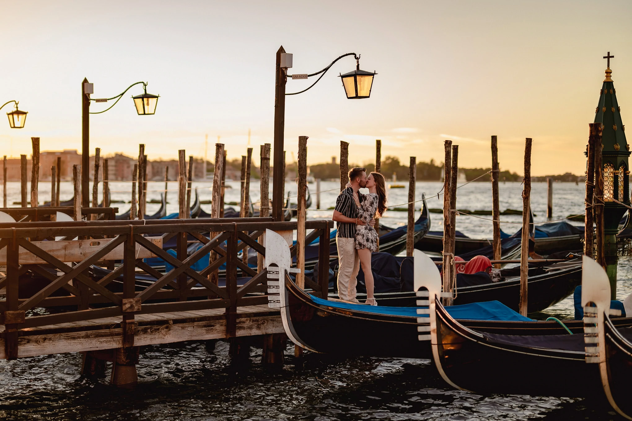 A couple shares a romantic kiss on a Venice dock as the sun sets, capturing a heartfelt proposal moment by the gondolas.