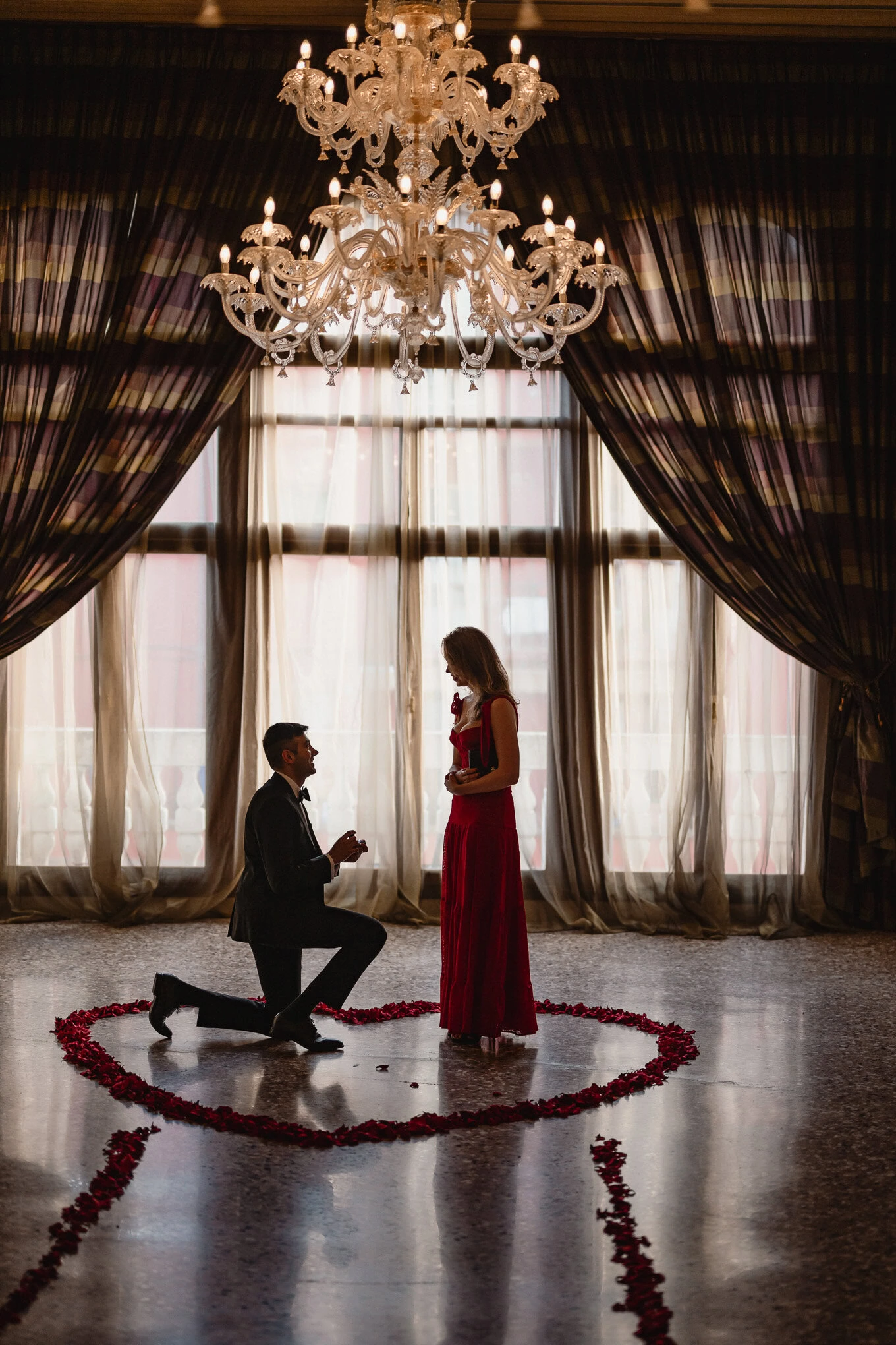 A man proposes to a woman inside an elegant room with a chandelier, surrounded by rose petals forming a heart shape.
