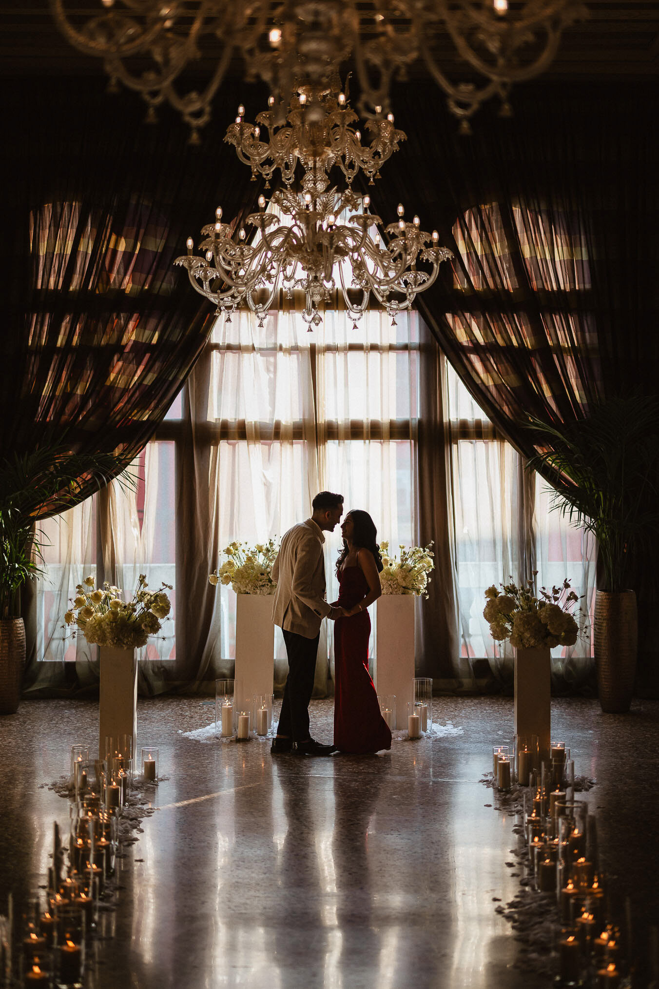 A couple standing in an elegant Venetian interior with a grand chandelier and large windows, creating a romantic atmosph.