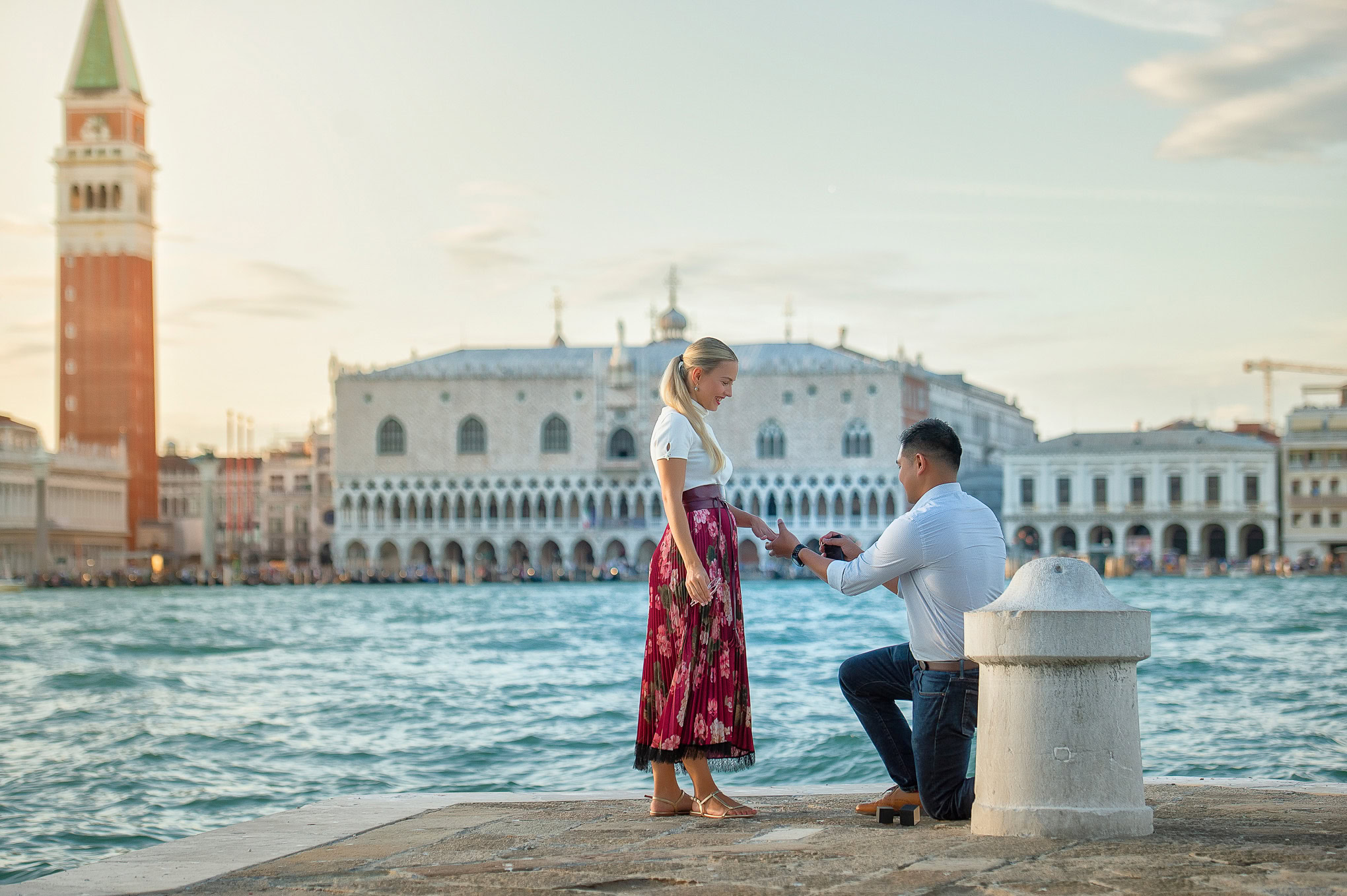 A woman reacts emotionally as a man kneels proposing on the Venice waterfront at sunset.