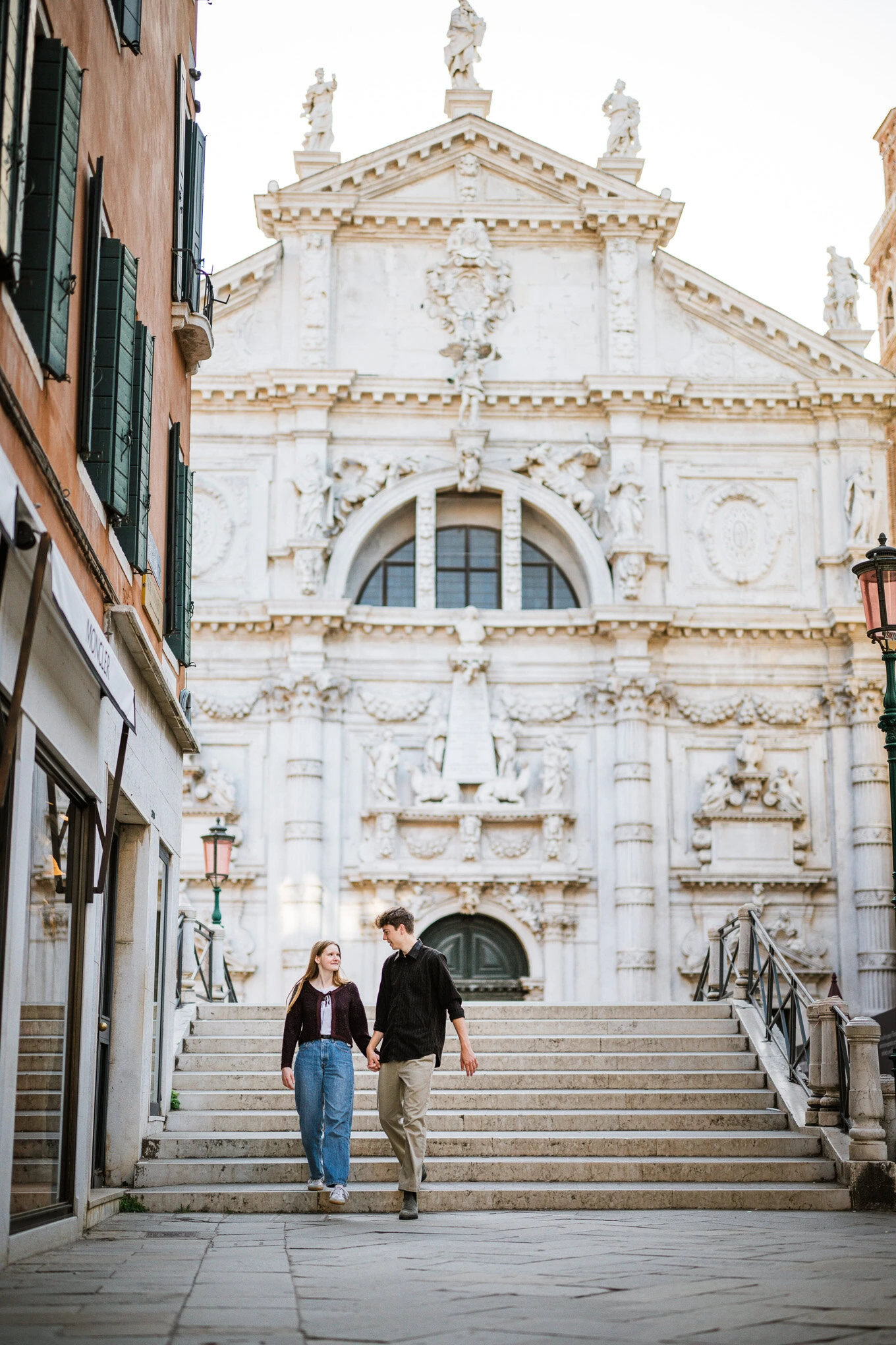 A couple holding hands and gazing at each other as they walk up the steps in front of a grand Venetian church.
