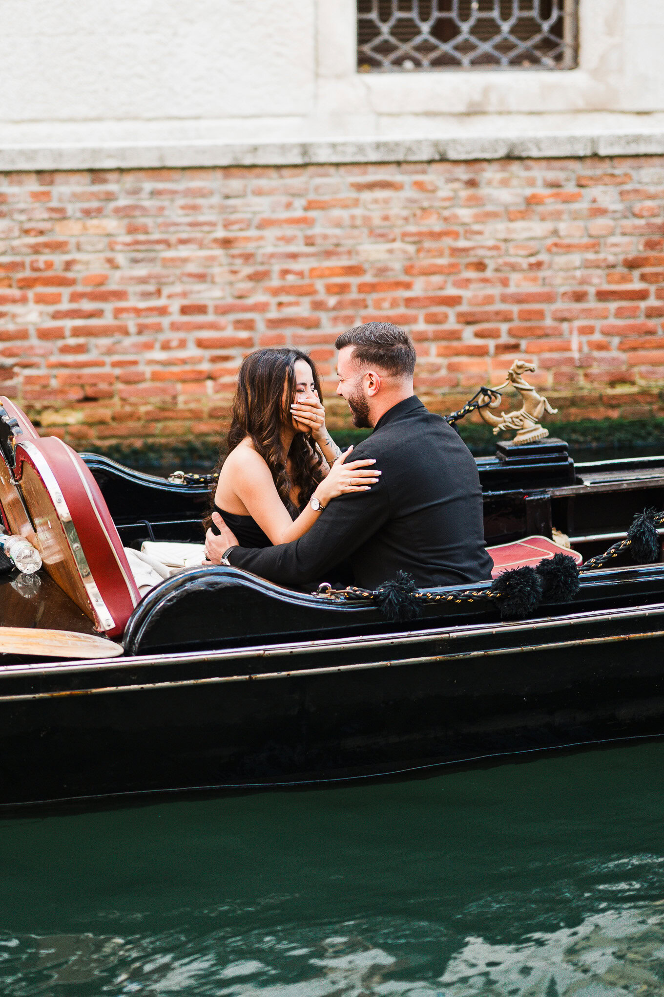 A couple sharing a romantic moment on a gondola in Venice, with historic brick walls in the background.