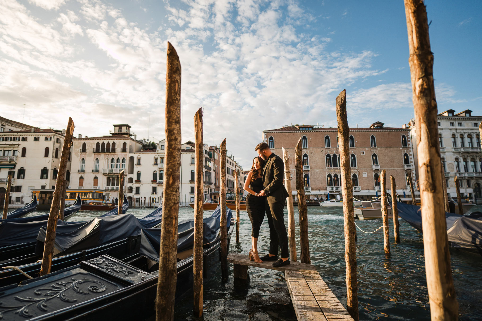 A couple shares a romantic proposal moment on a Venice dock, surrounded by gondolas and historic buildings under a brigh.