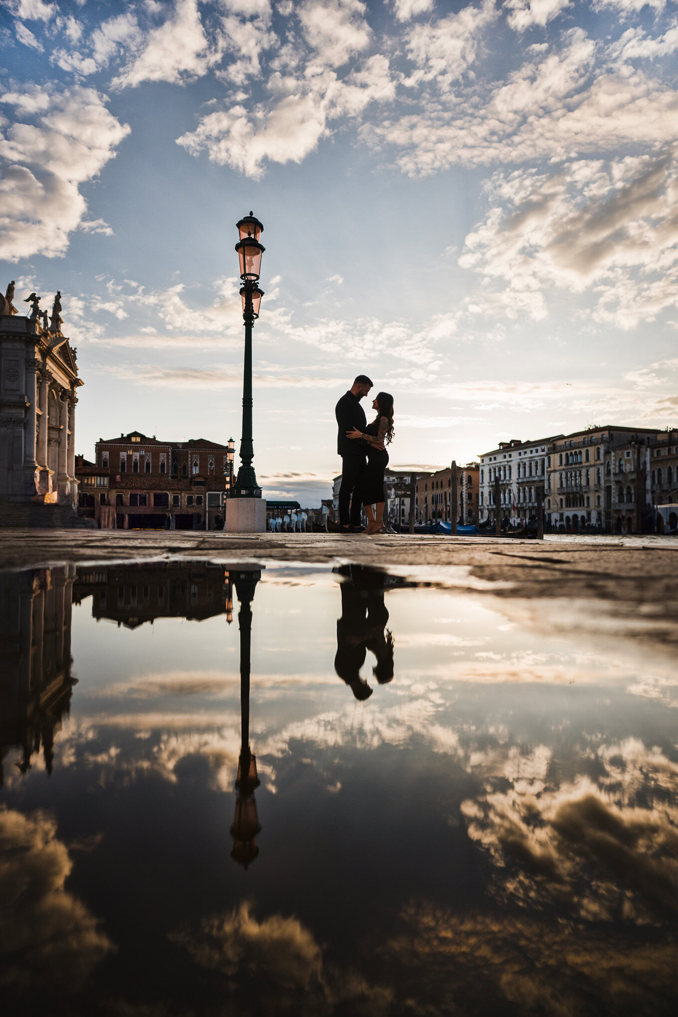 A couple embracing near a Venetian canal under a dramatic sky, captured in a moody, atmospheric style.