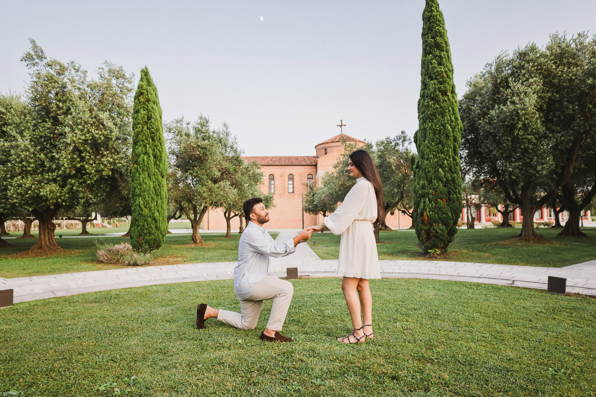 A man proposing to a woman in a lush Venetian park with tall cypress trees and historic buildings in the background.
