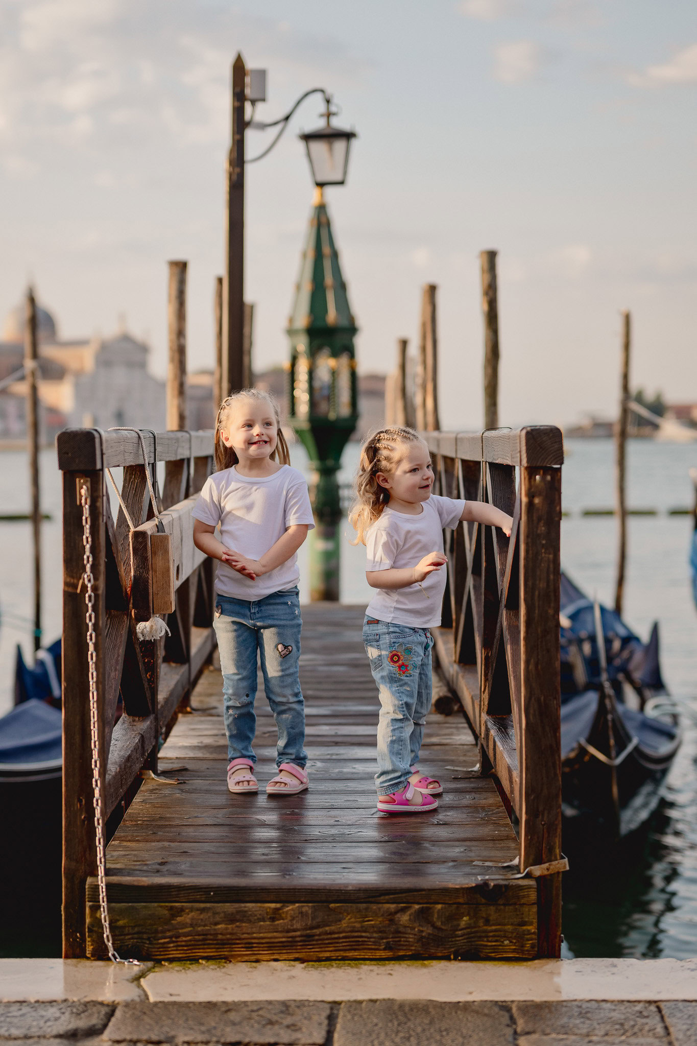 Children standing on a wooden dock near water with boats and historic buildings in the background.