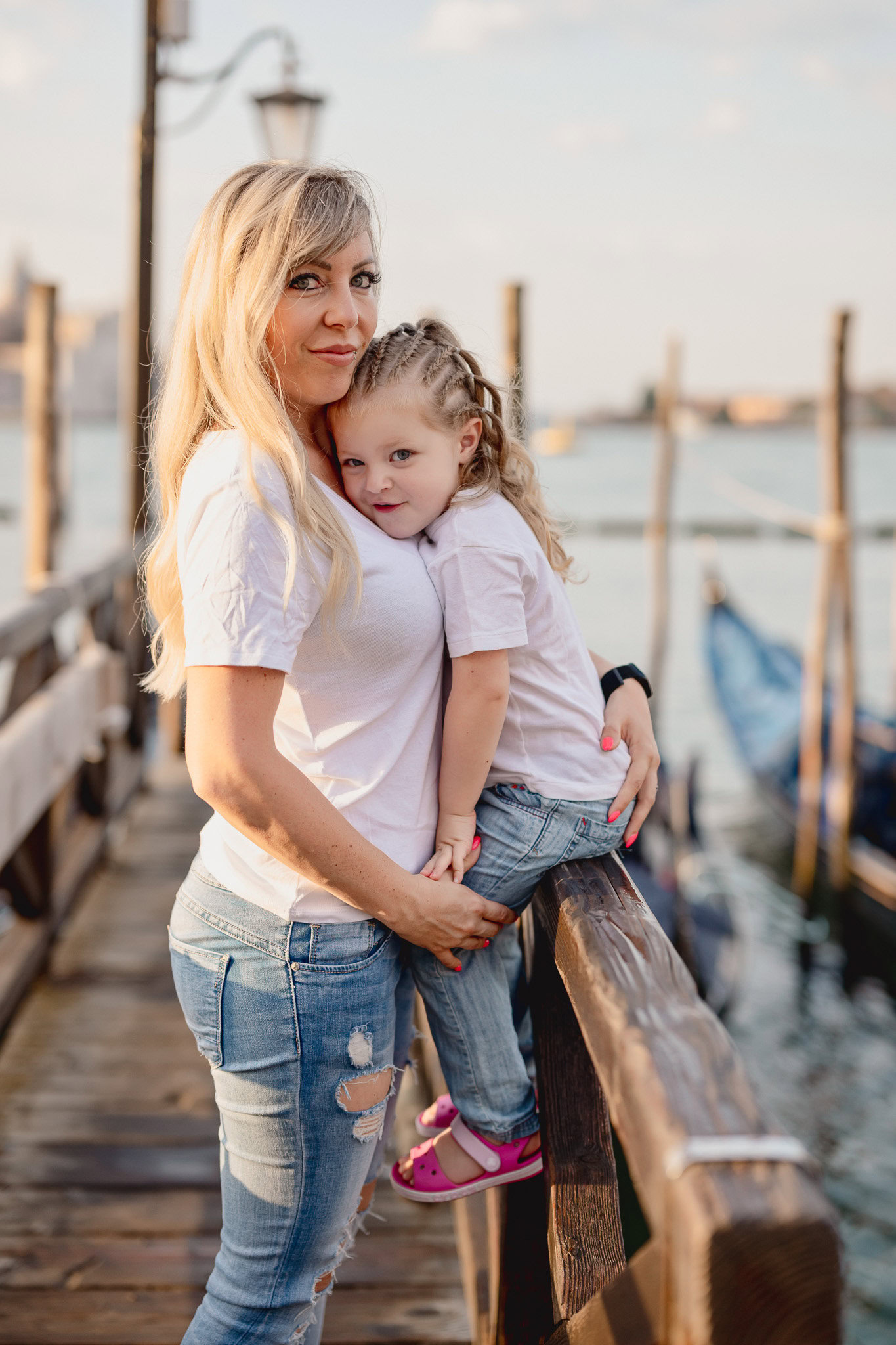 A woman and a young girl standing on a wooden dock near water with boats in the background.