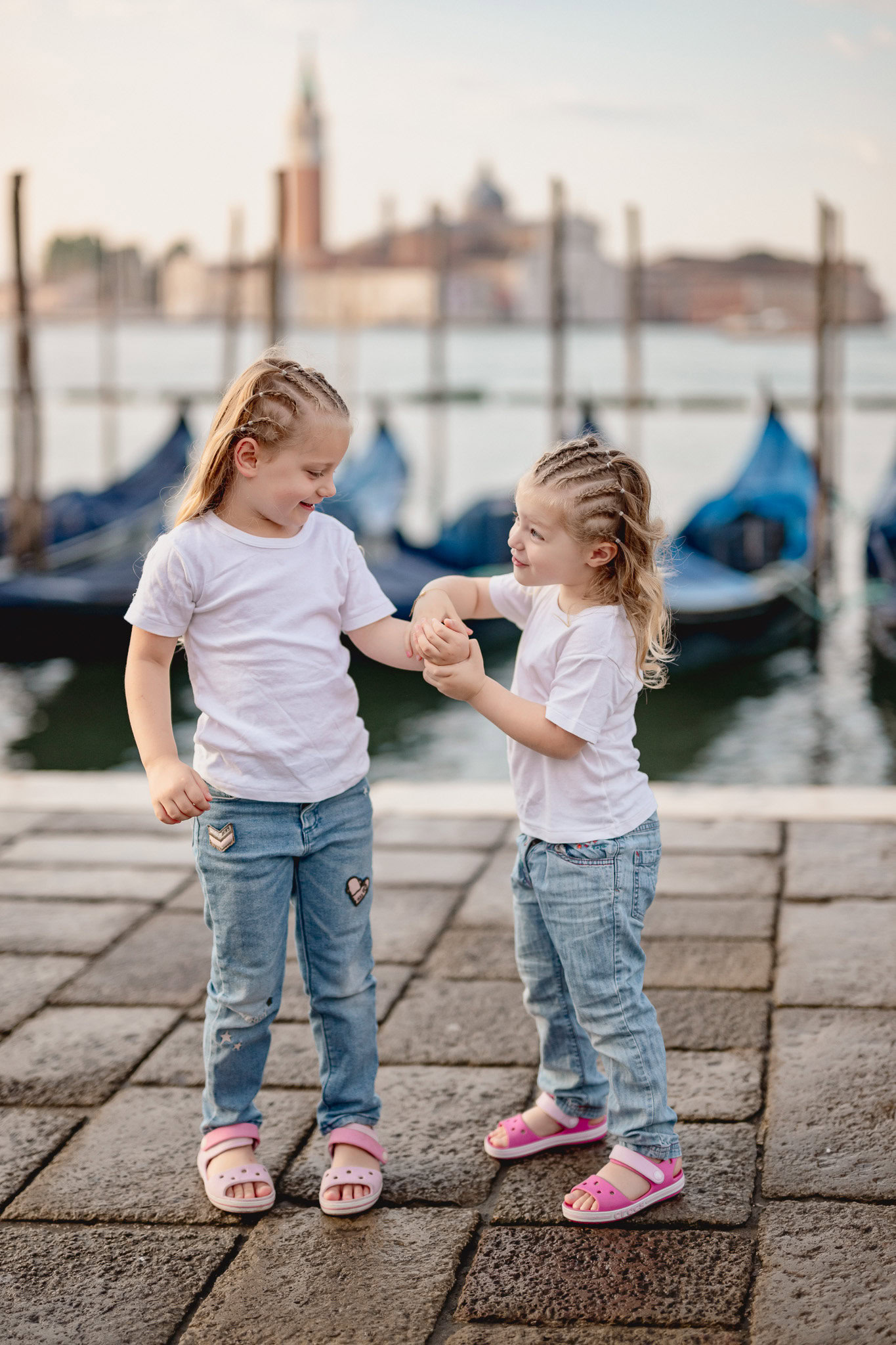 A girl and a younger girl holding hands near a canal with boats and historic buildings in the backgr.