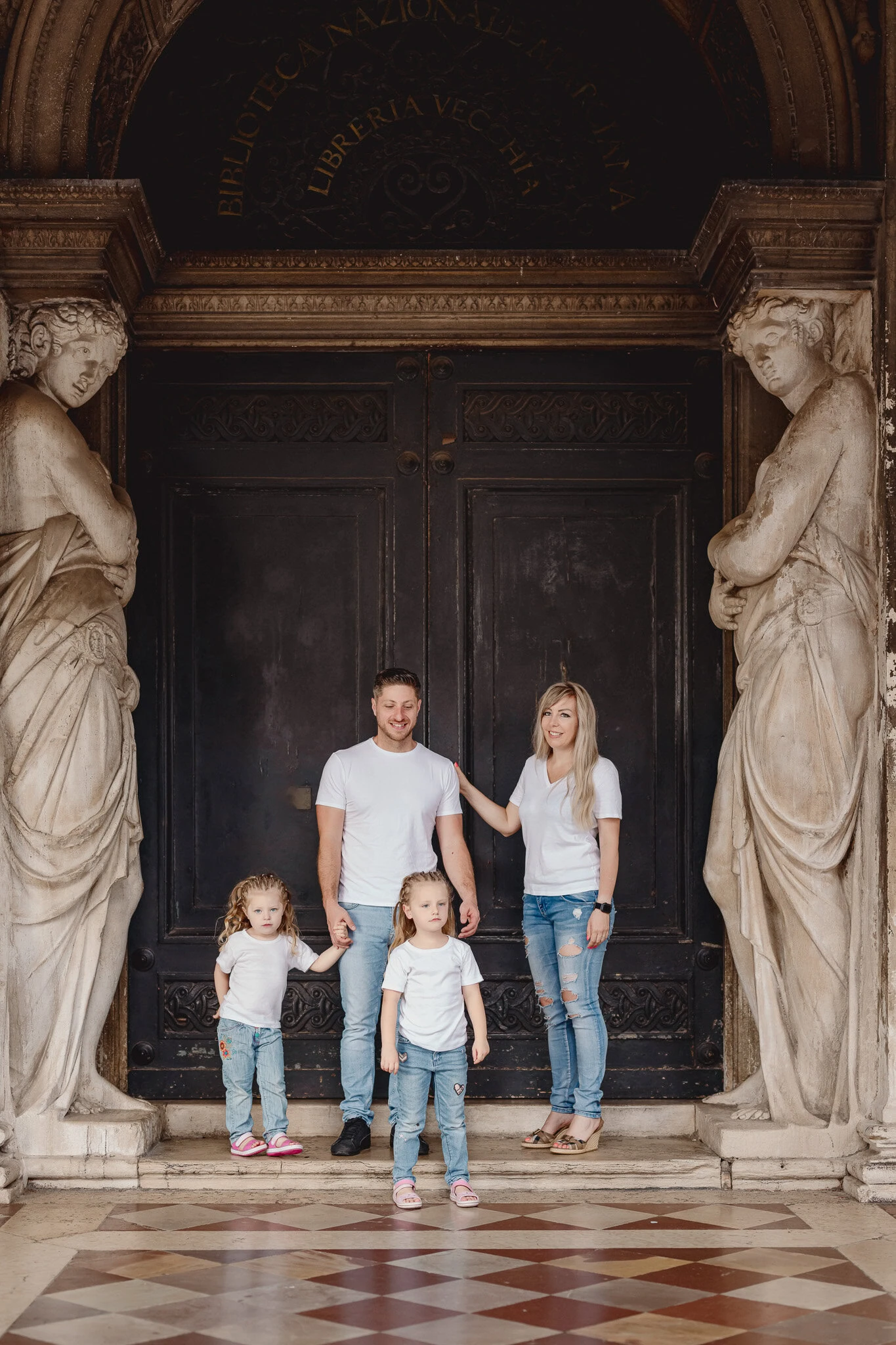 A woman, man, and two young girls standing in front of a large ornate door flanked by classical stat.