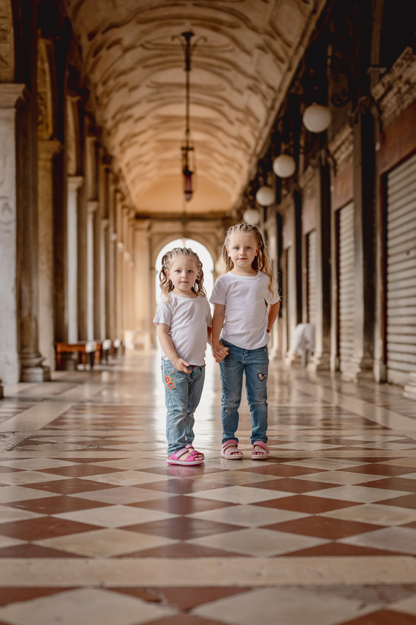 Children standing in an arched corridor with patterned flooring and hanging lights, with shops and b.
