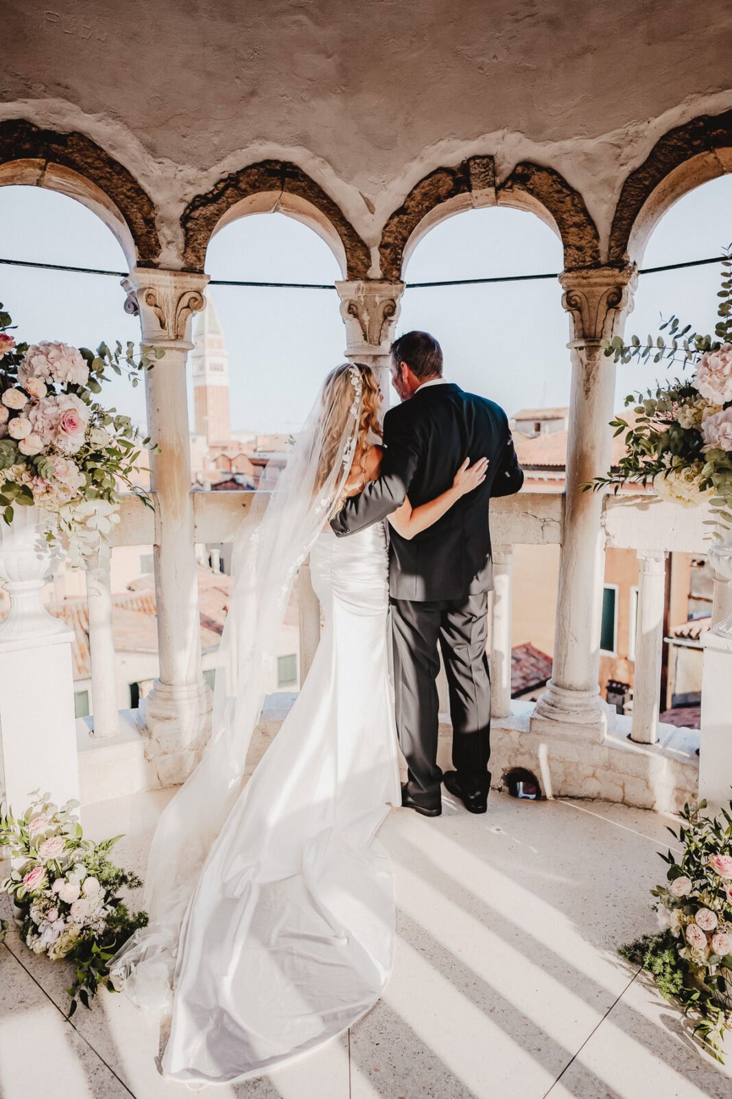 Elegant couple standing on a balcony with floral arrangements and historic architecture in the backg.