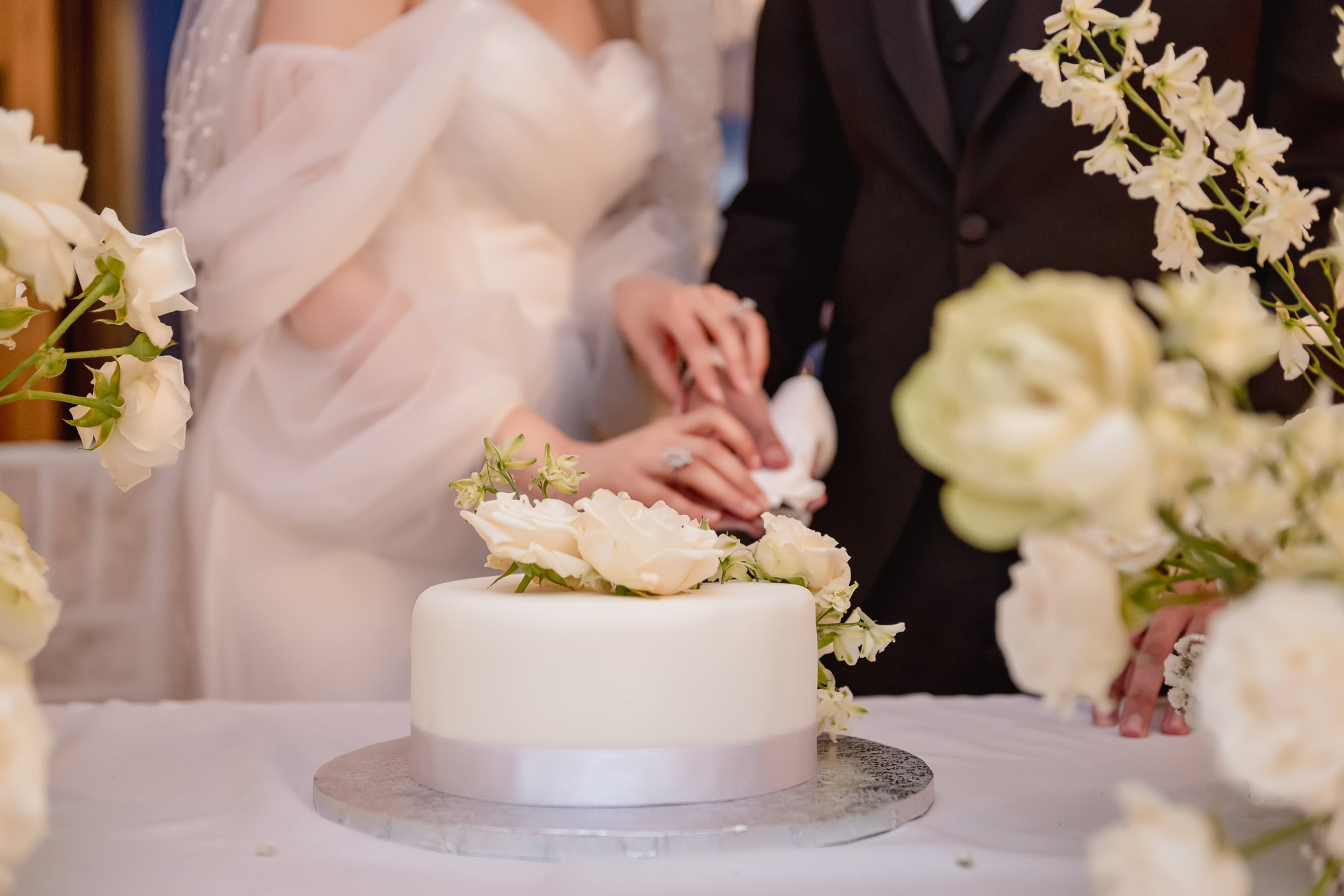A couple's hands gently clasped over a white wedding cake adorned with soft white flowers, set against a romantic Veneti.