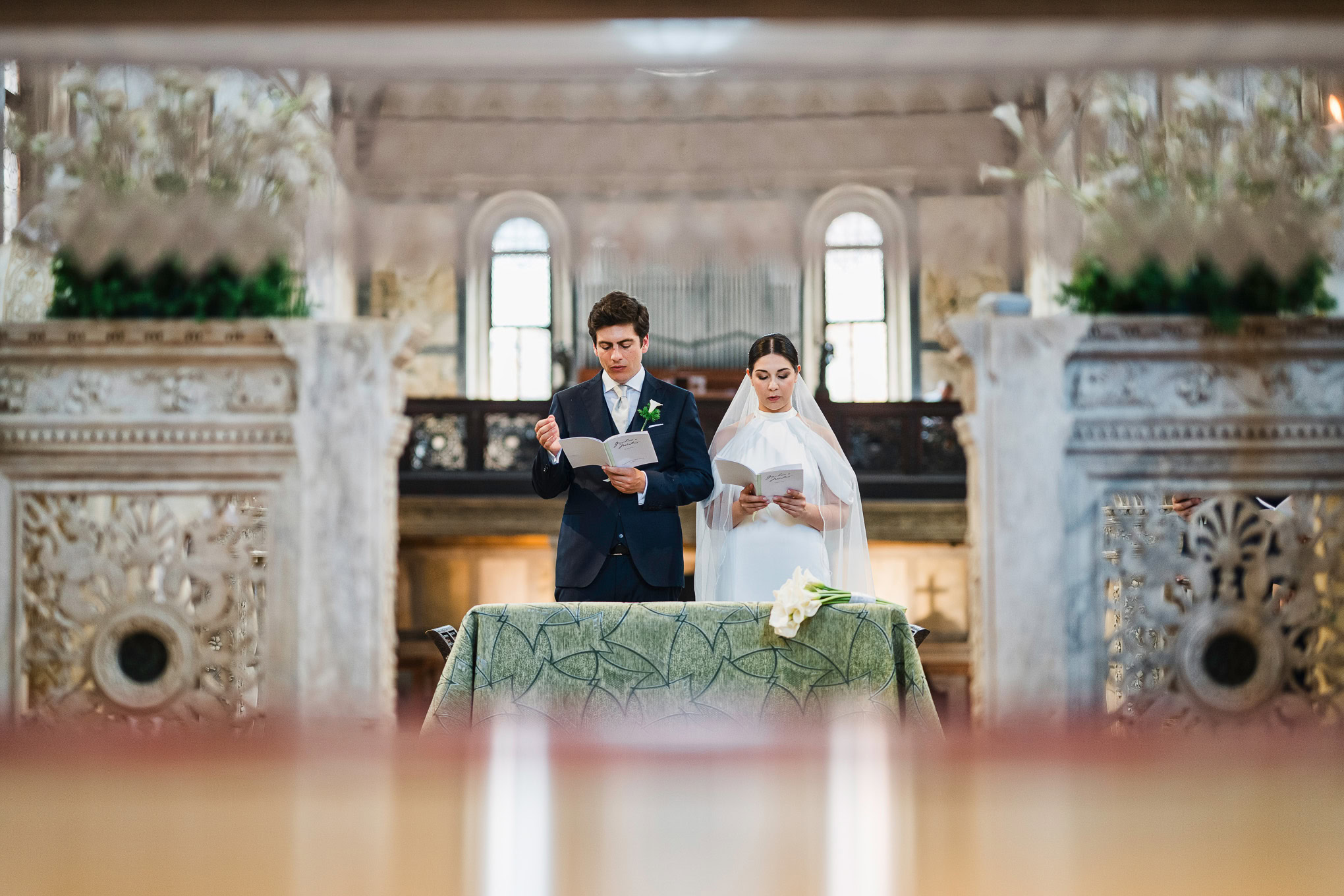 A couple exchanging vows during a wedding ceremony inside a historic Venetian church, illuminated by soft natural light.