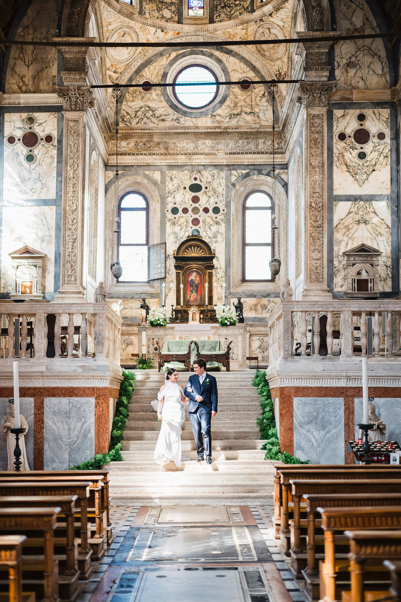 A couple shares a tender moment at their wedding inside a grand, sunlit Venetian church with ornate marble walls and his.