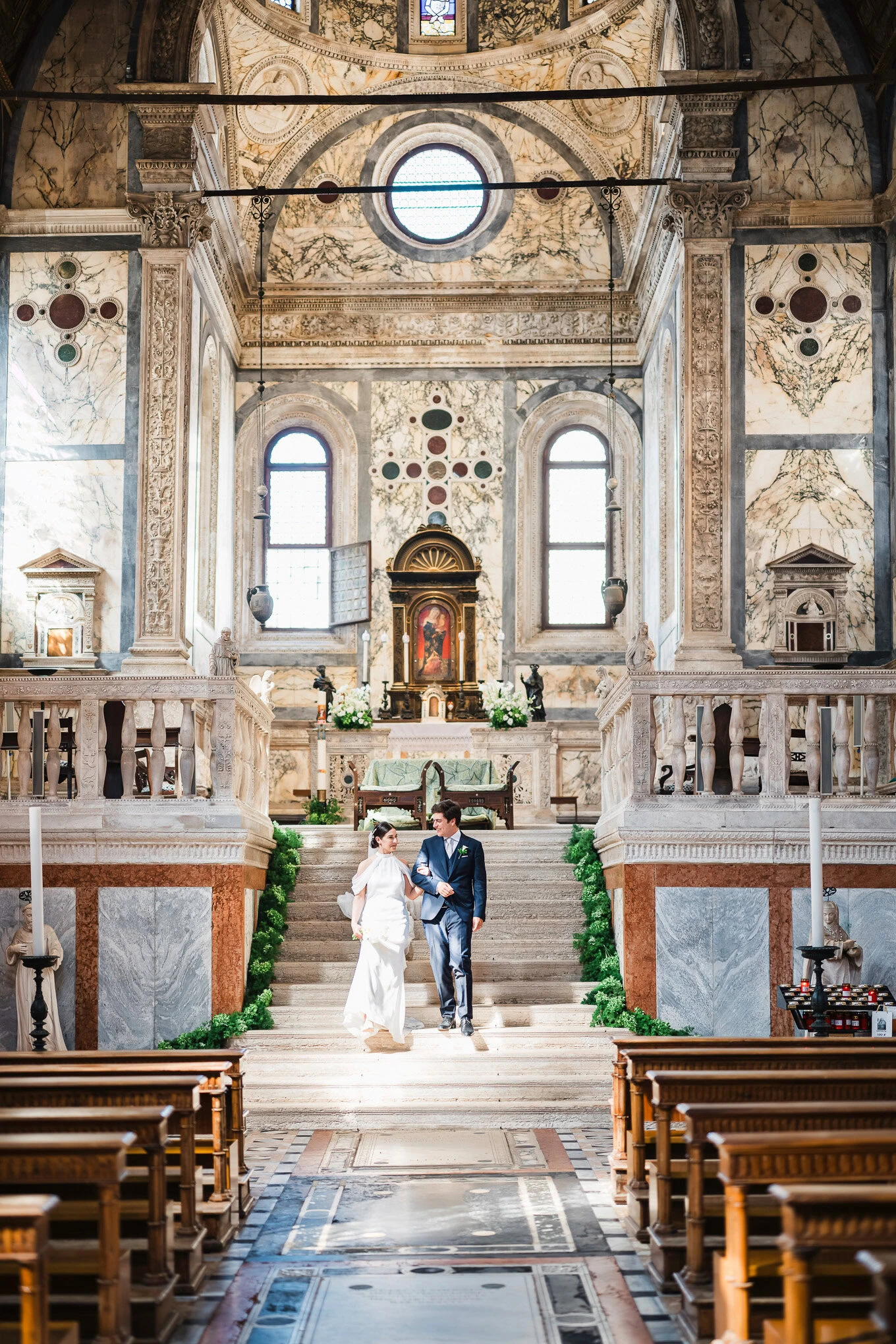 A couple shares a tender moment at their wedding inside a grand, sunlit Venetian church with ornate marble walls and his.