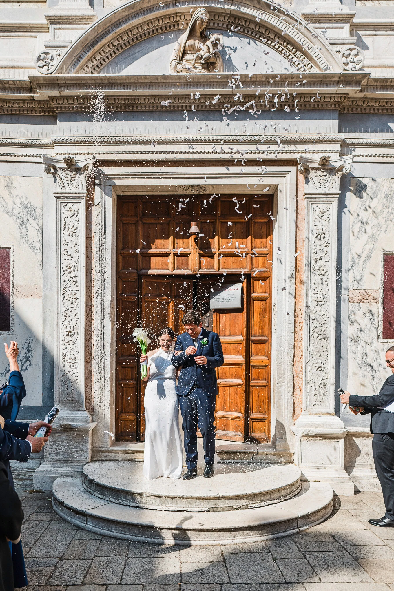 Elegant newlyweds celebrating their wedding outside a historic Venetian church, sunlight illuminating their joyful momen.