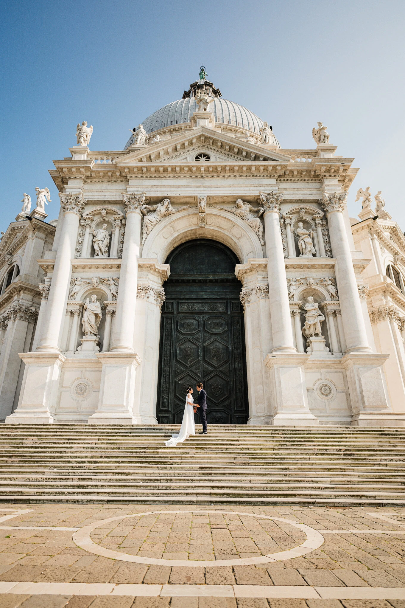 A stunning wedding couple standing on the grand steps of a historic Venetian church under a clear blue sky, illuminated.