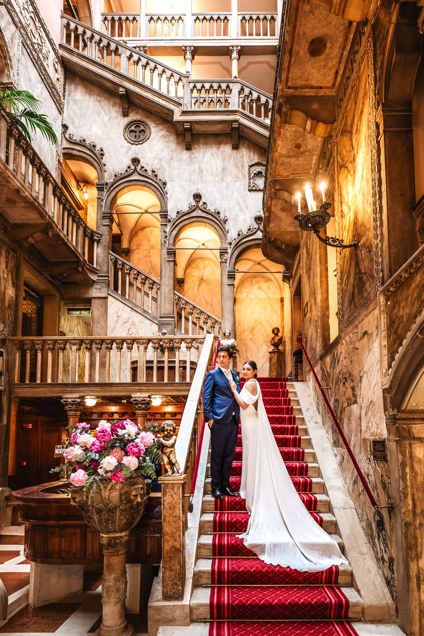 Elegant bride and groom standing on a grand Venetian staircase illuminated by warm, ambient lighting in a historic palac.