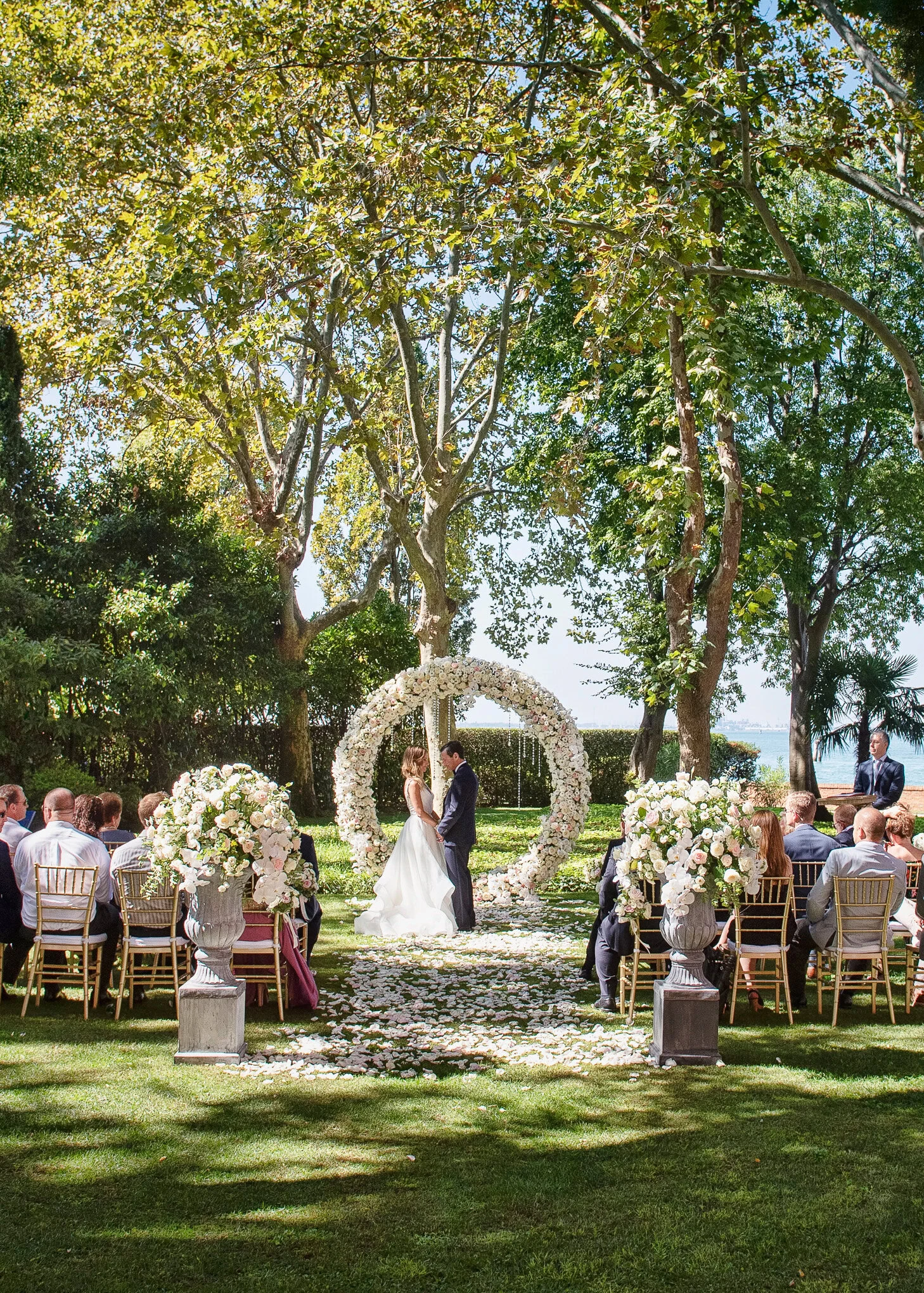 A lush outdoor wedding ceremony in Venice, with a romantic couple exchanging vows beneath a floral arch, surrounded by t.