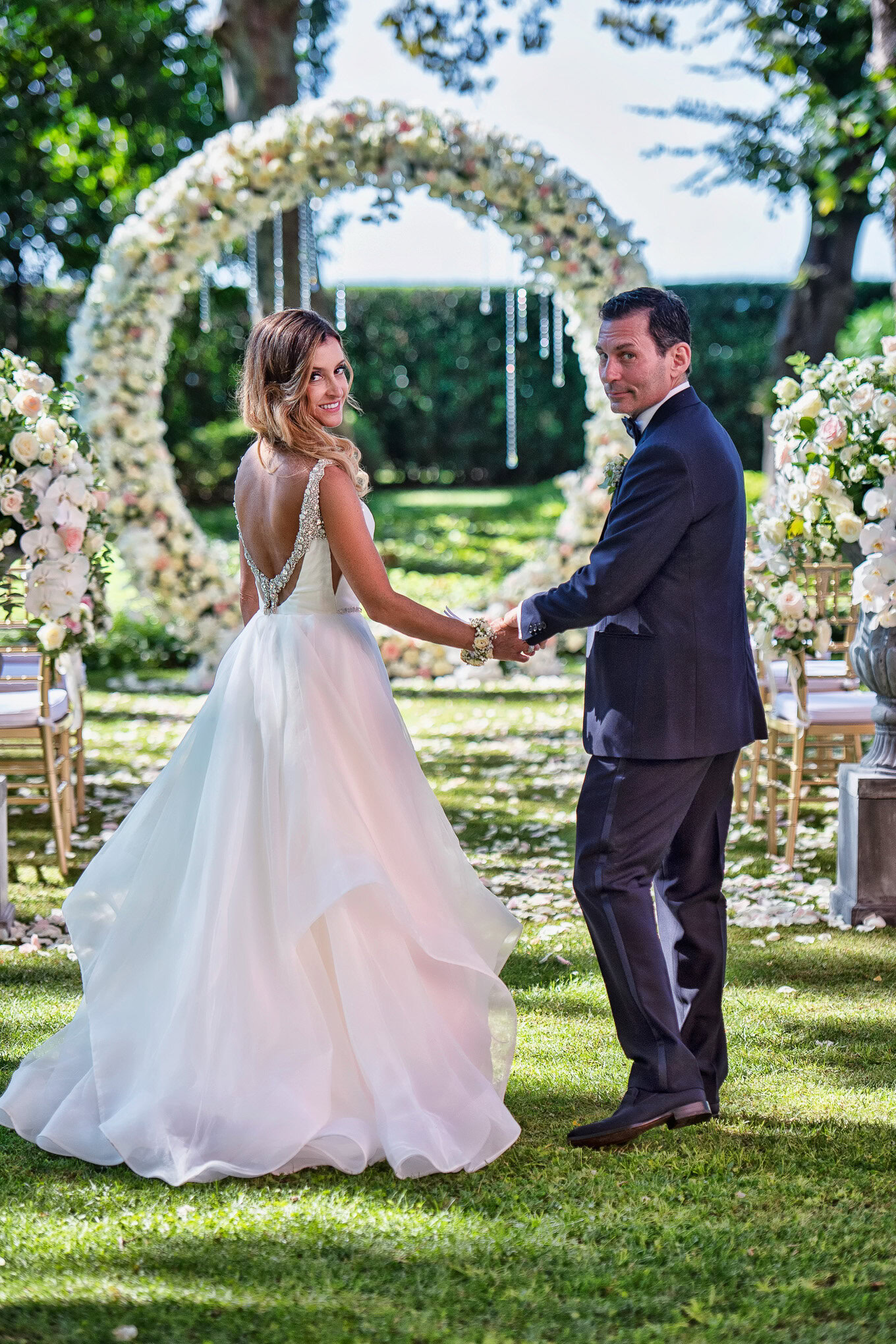 A couple holding hands during a wedding ceremony in a lush Venetian garden with floral decorations.