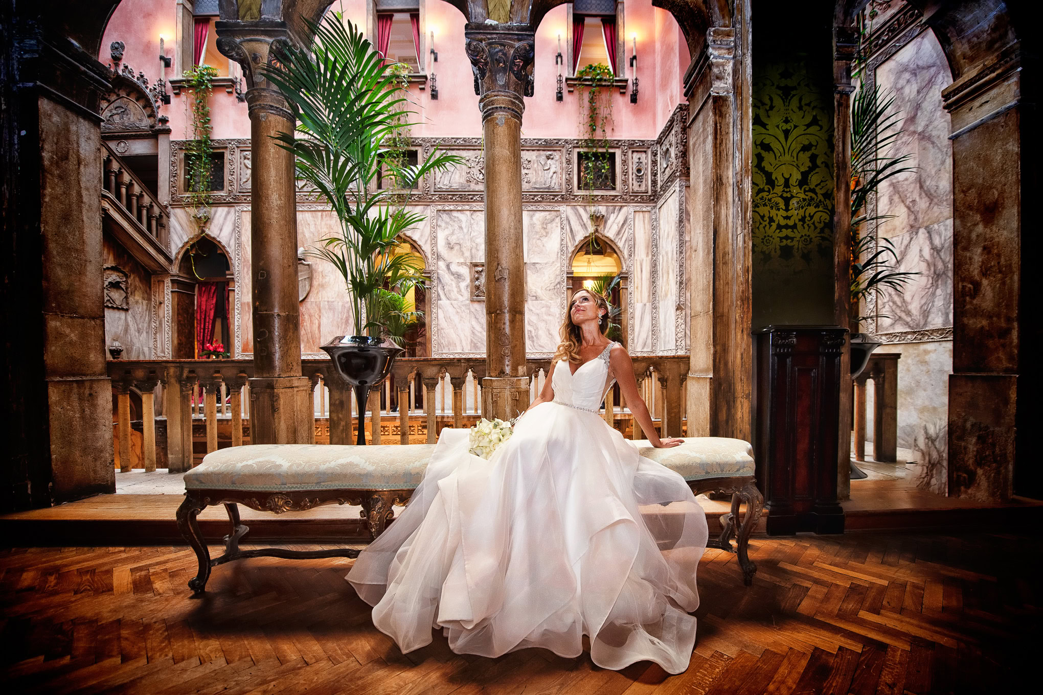 A woman in a wedding dress sitting on a vintage bench inside an ornate Venetian-style interior.