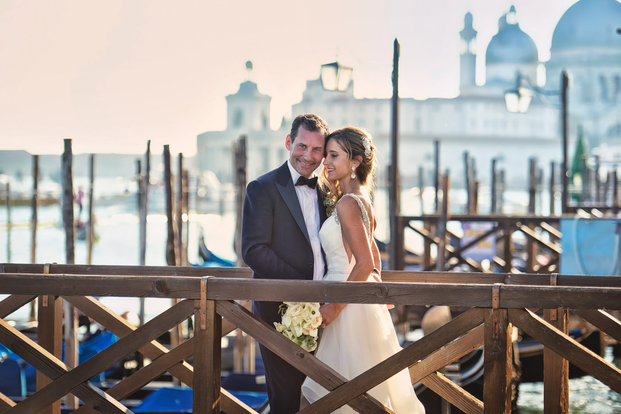 A couple in wedding attire sharing a joyful moment on a Venice dock with gondolas and historic architecture in the backg.