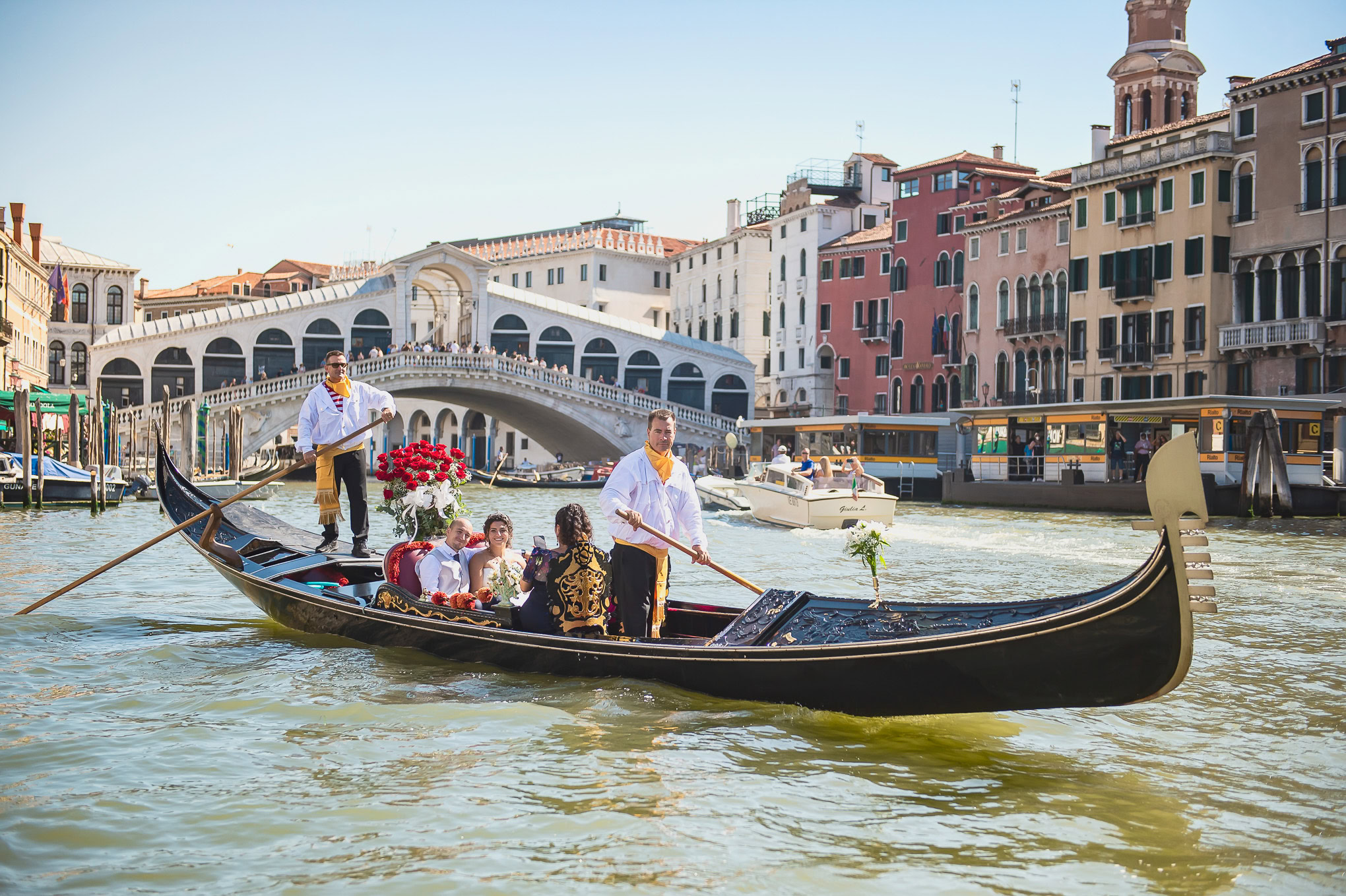 A gondola glides through the shimmering Venetian canals as a joyful wedding celebration unfolds against colorful histori.